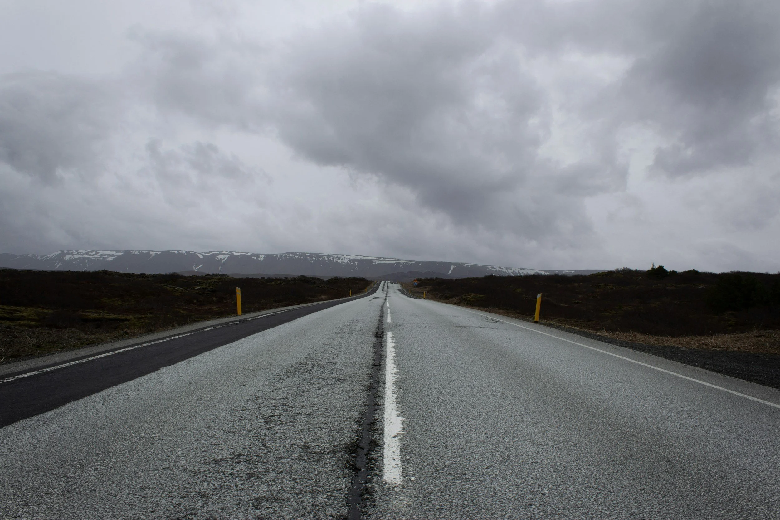 A long empty road stretching into the distance under a cloudy, overcast sky with snow-capped mountains in the background.