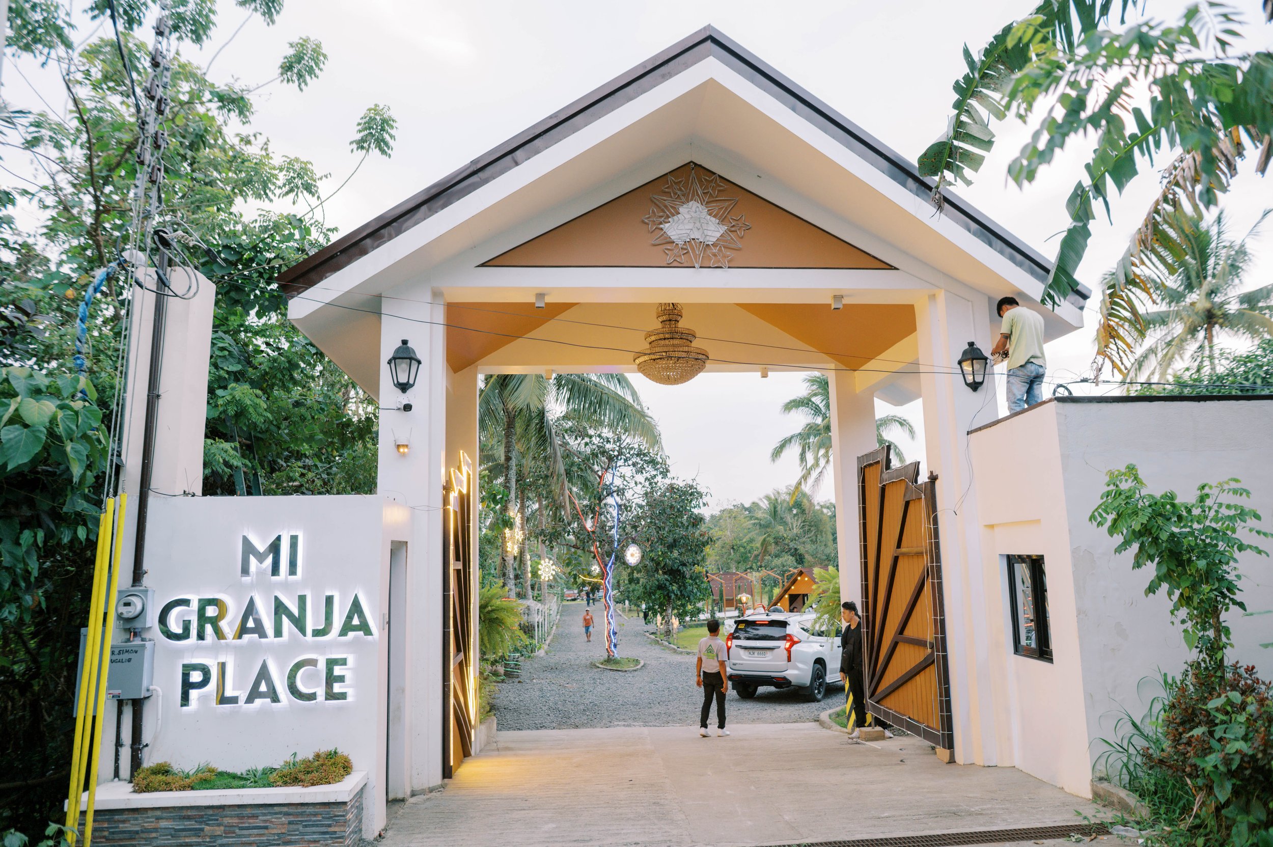 Entrance gate to a property called 'Mi Granja Place' with decorative lighting, palm trees, and a gravel pathway leading inside.