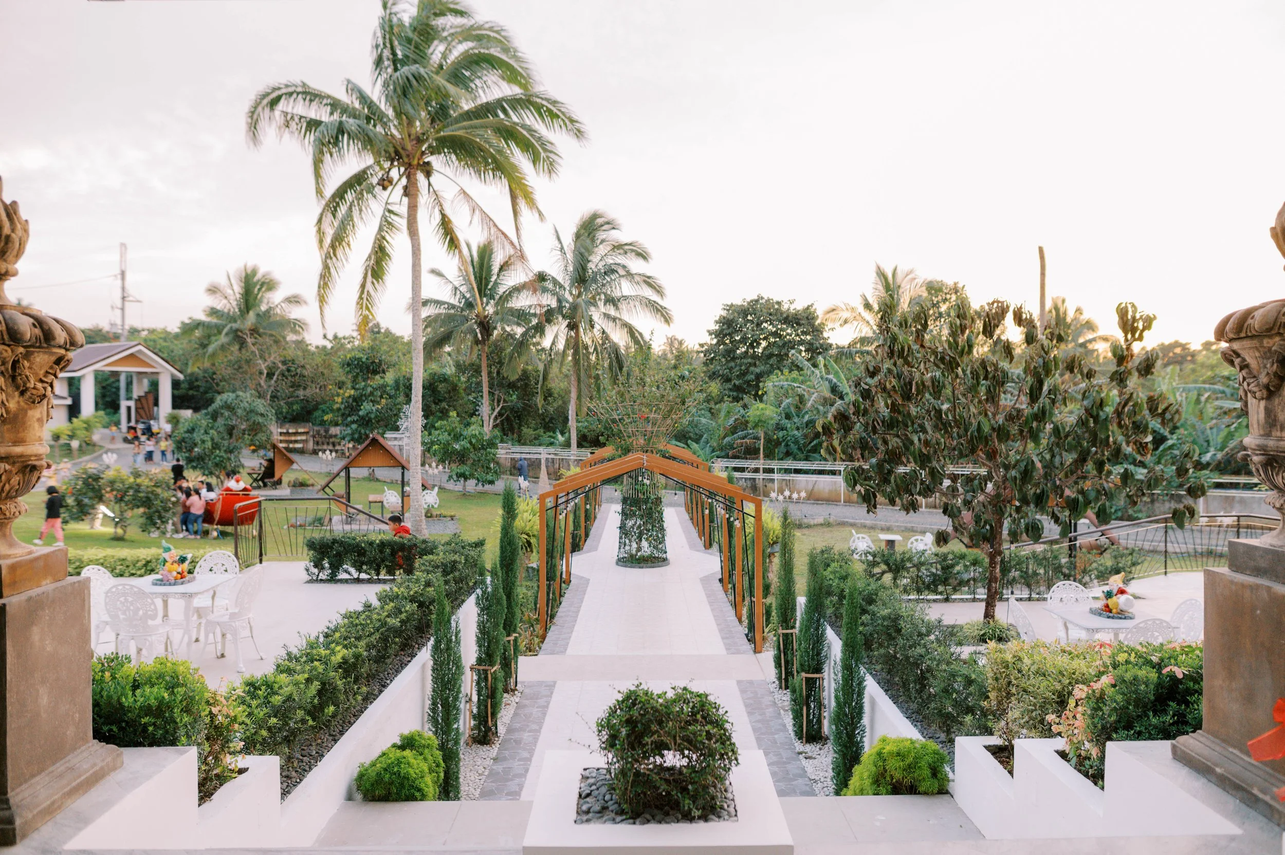 View of a garden with palm trees, pathways, benches, and people in the background, taken from a staircase or balcony.