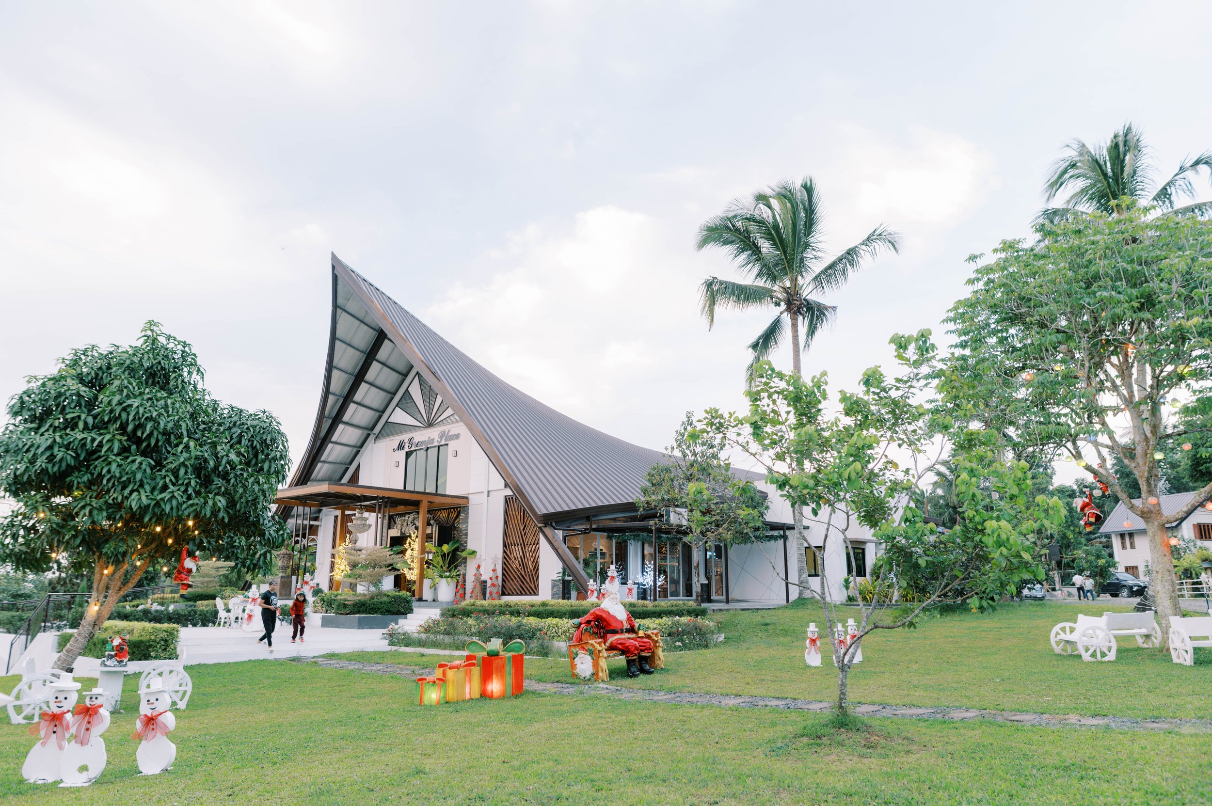 A modern building with an A-frame roof, surrounded by trees and holiday decorations, including snowmen, Santa figures, and gift boxes, on a grassy lawn during daytime.