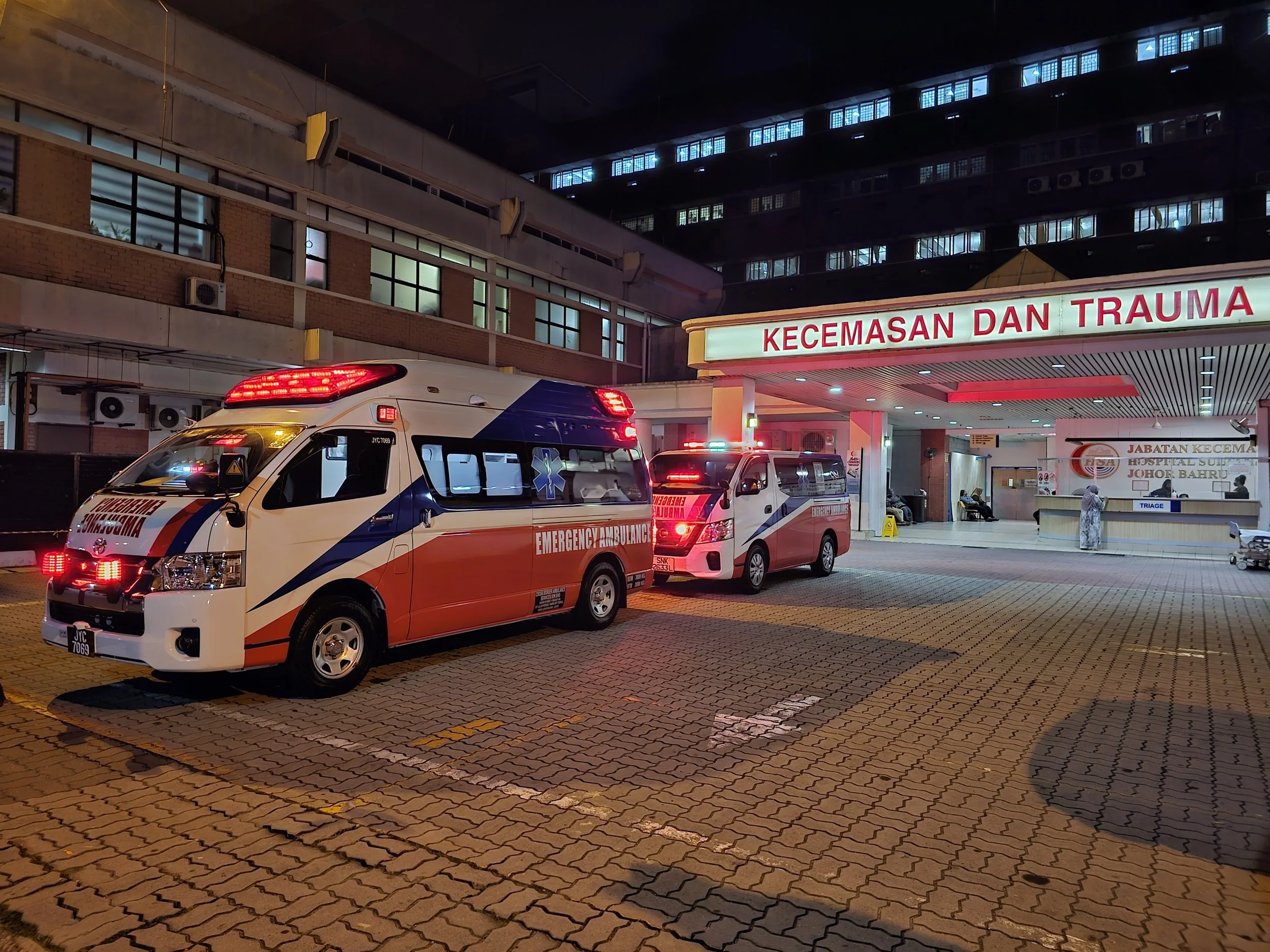 Emergency ambulances parked outside the emergency and trauma department of a hospital at night, with hospital signage and a person standing near the entrance.