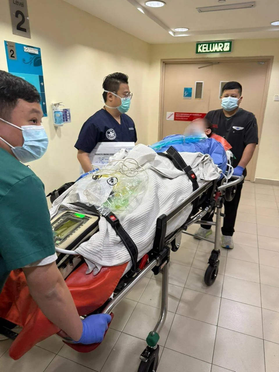 Medical team attending to a patient on a stretcher in a hospital corridor, with all individuals wearing masks.