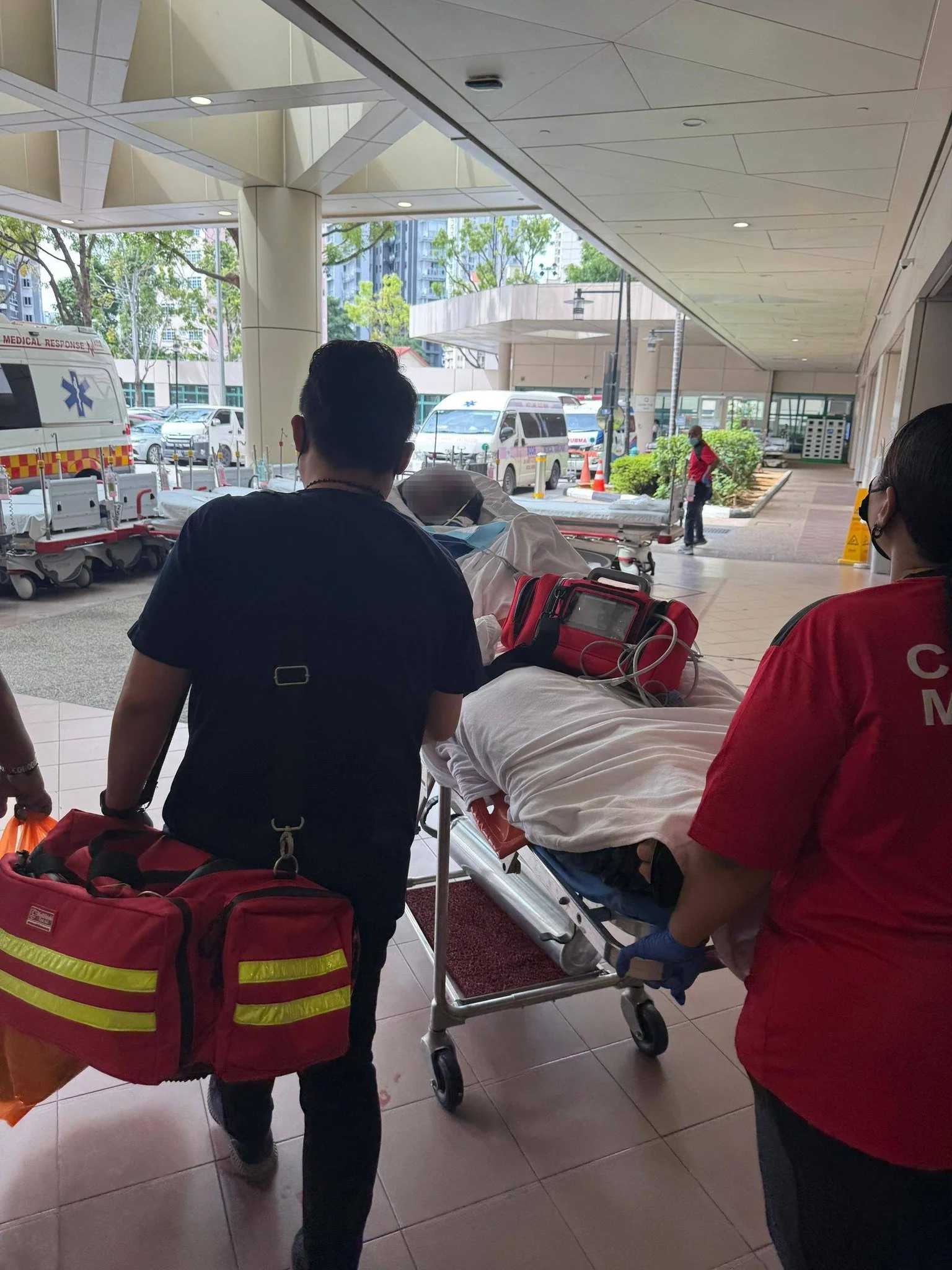 Medical emergency personnel transporting a patient on a stretcher into a hospital, with ambulances and medical equipment visible outside.