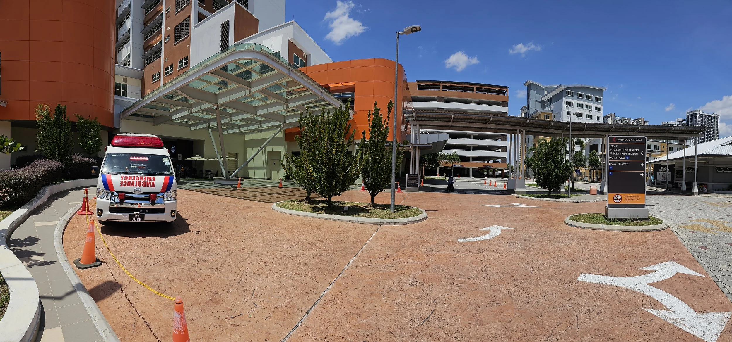 An ambulance parked near the entrance of a hospital, surrounded by orange traffic cones and pedestrian pathways, with a clear blue sky overhead and modern buildings in the background.