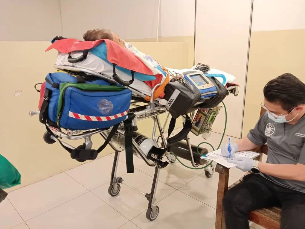 A patient lying on a stretcher connected to medical devices in a hospital, with a healthcare worker taking notes.