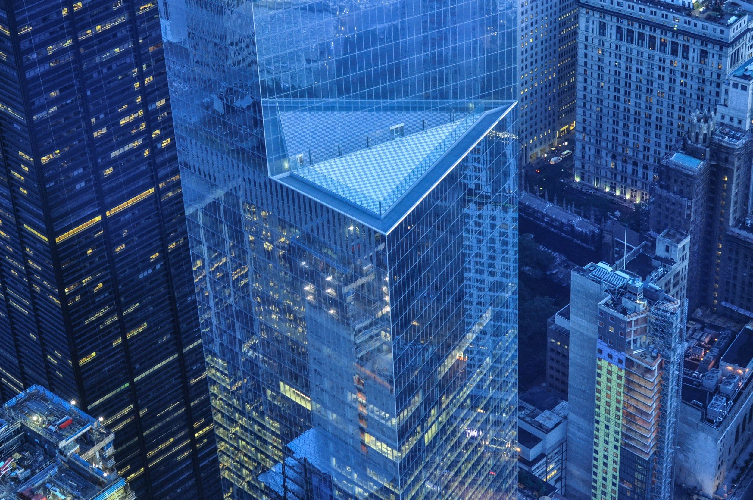 Nighttime aerial view of tall city skyscraper with glass exterior and a rooftop terrace, surrounded by other high-rise buildings in a metropolitan area.