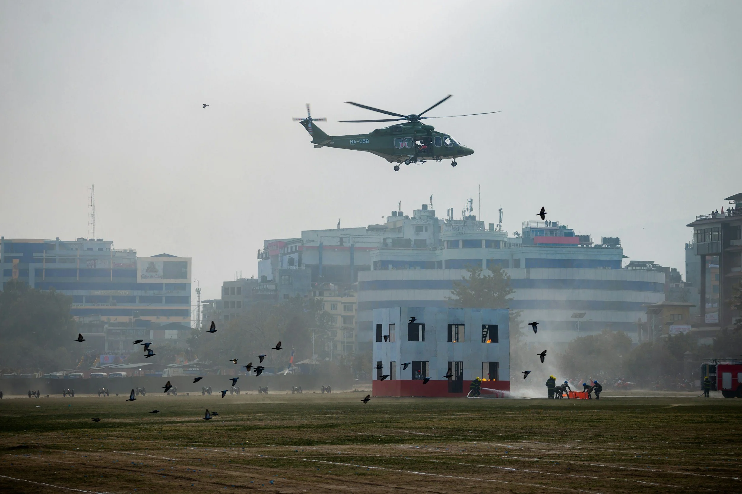 A helicopter flying over a cityscape with buildings, birds flying, and firefighters working on the ground, possibly putting out a fire.