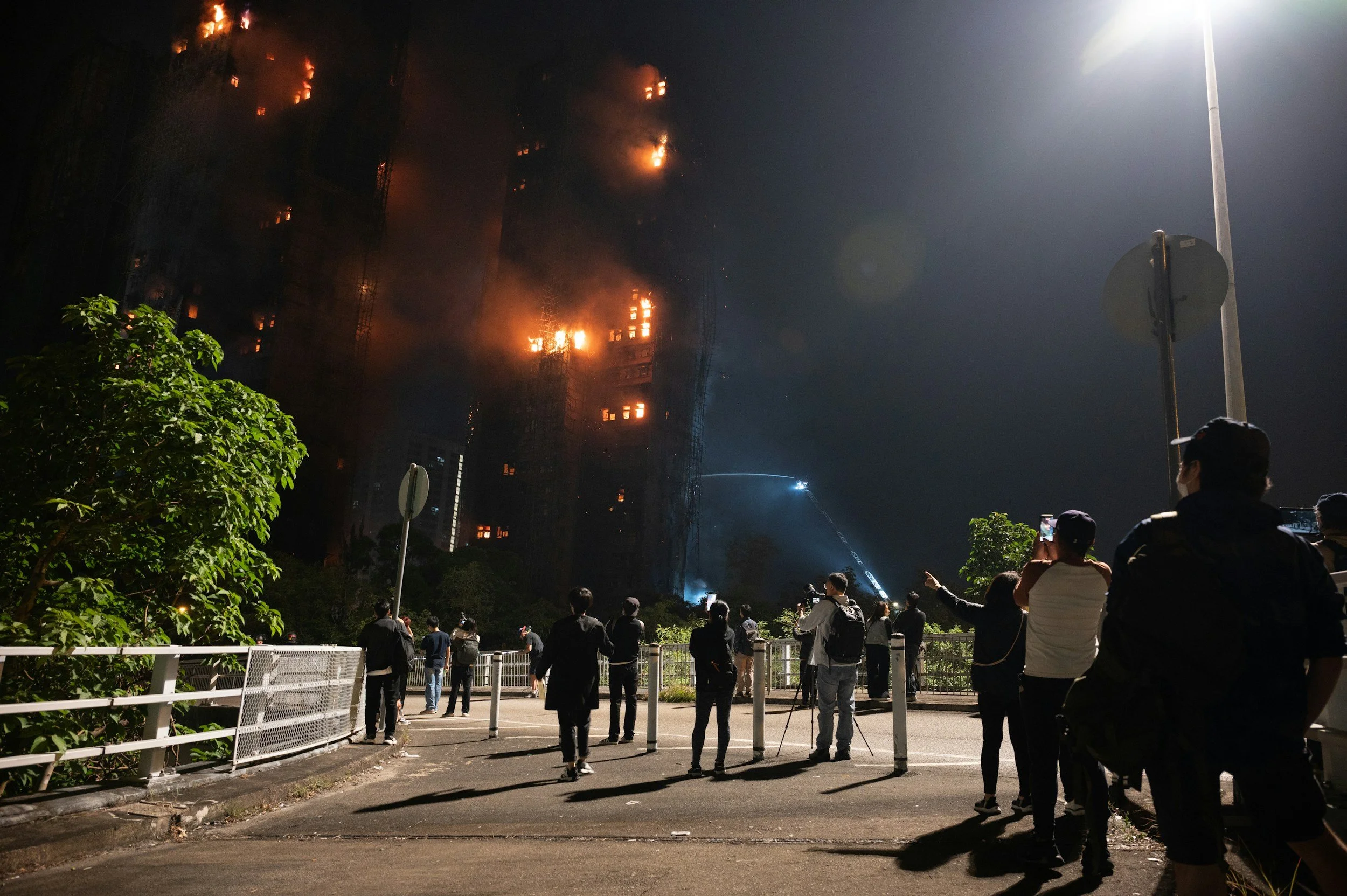 Nighttime scene of a high-rise building on fire with flames and smoke visible on multiple floors, while a crowd of people gather below, some taking photos or videos of the incident.