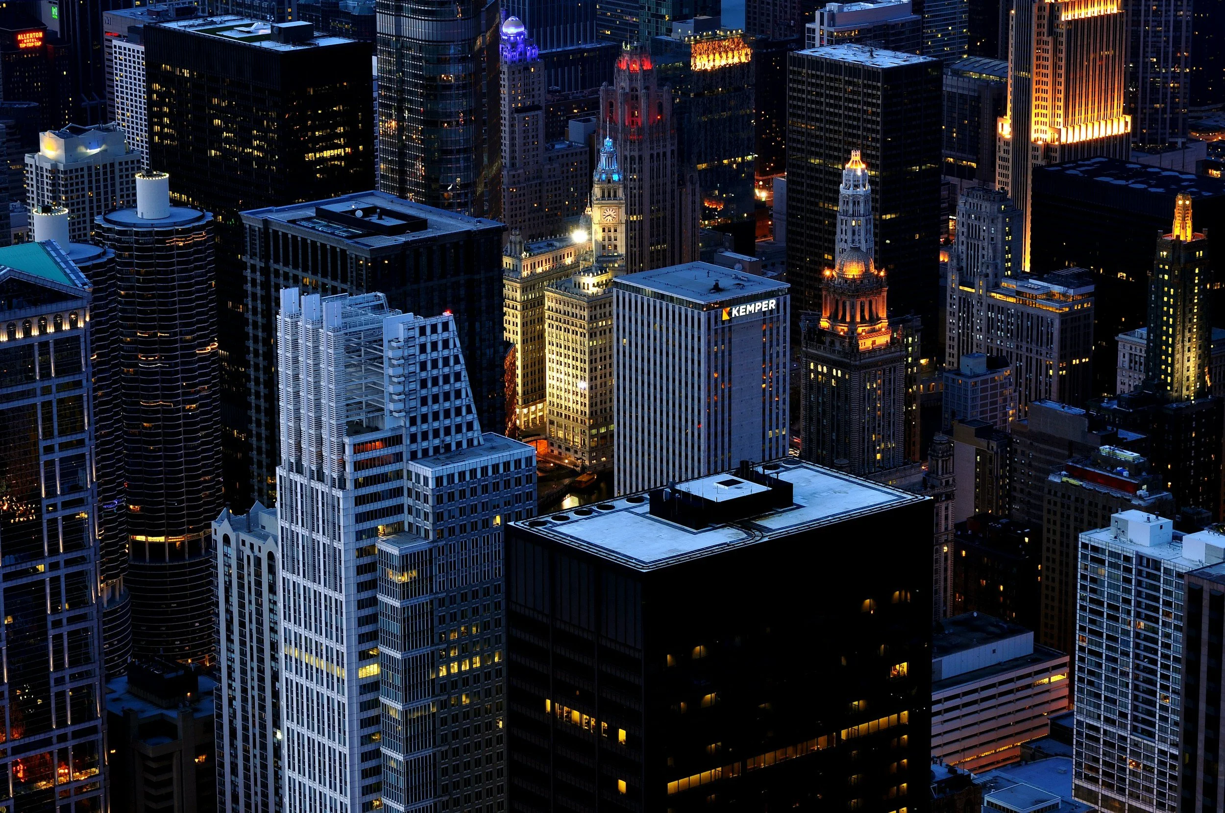 Nighttime cityscape of a downtown area with illuminated skyscrapers and historic buildings, viewed from above.