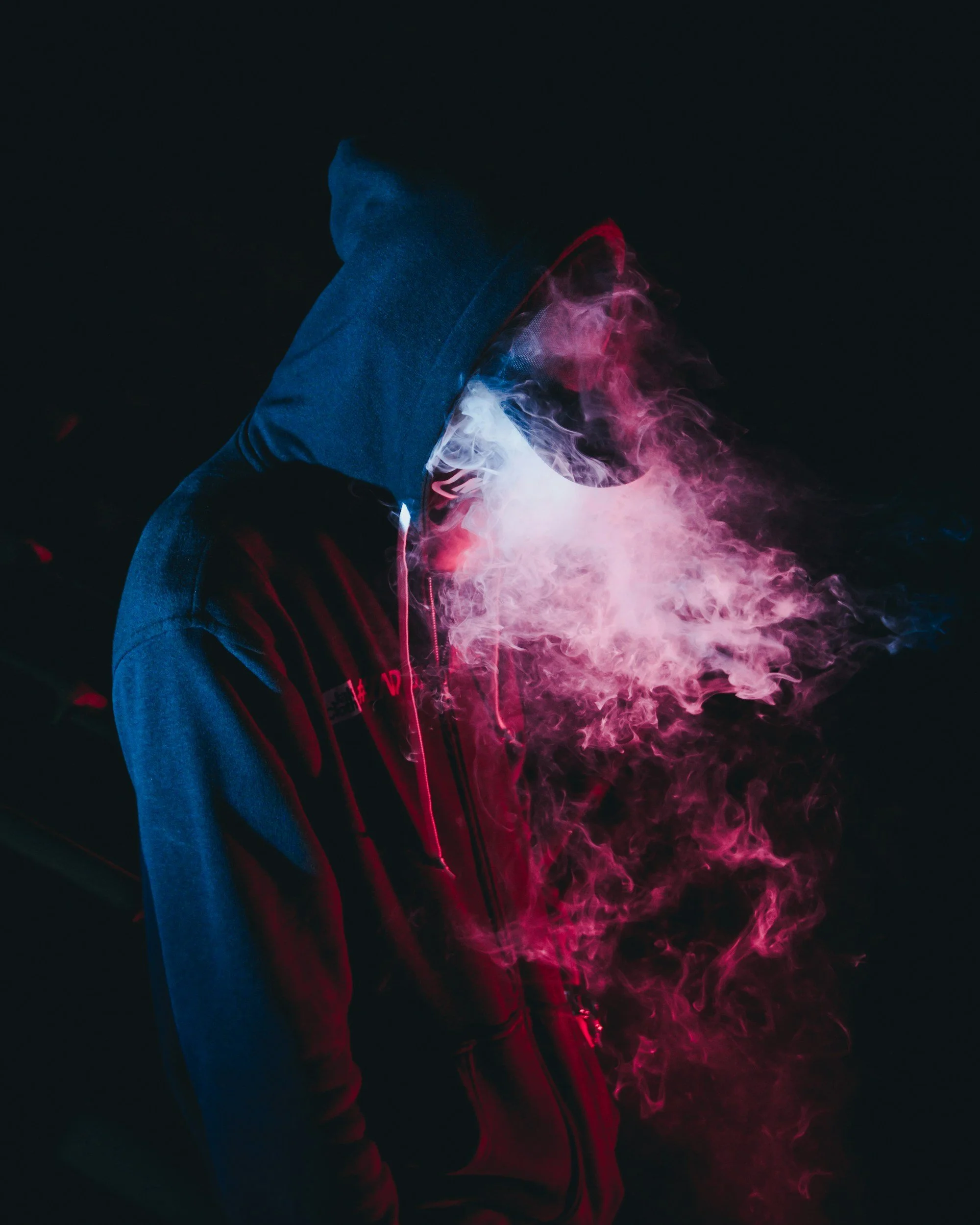 A person wearing a hoodie stands in front of a dark background, illuminated by colorful smoke in shades of pink, white, and purple.