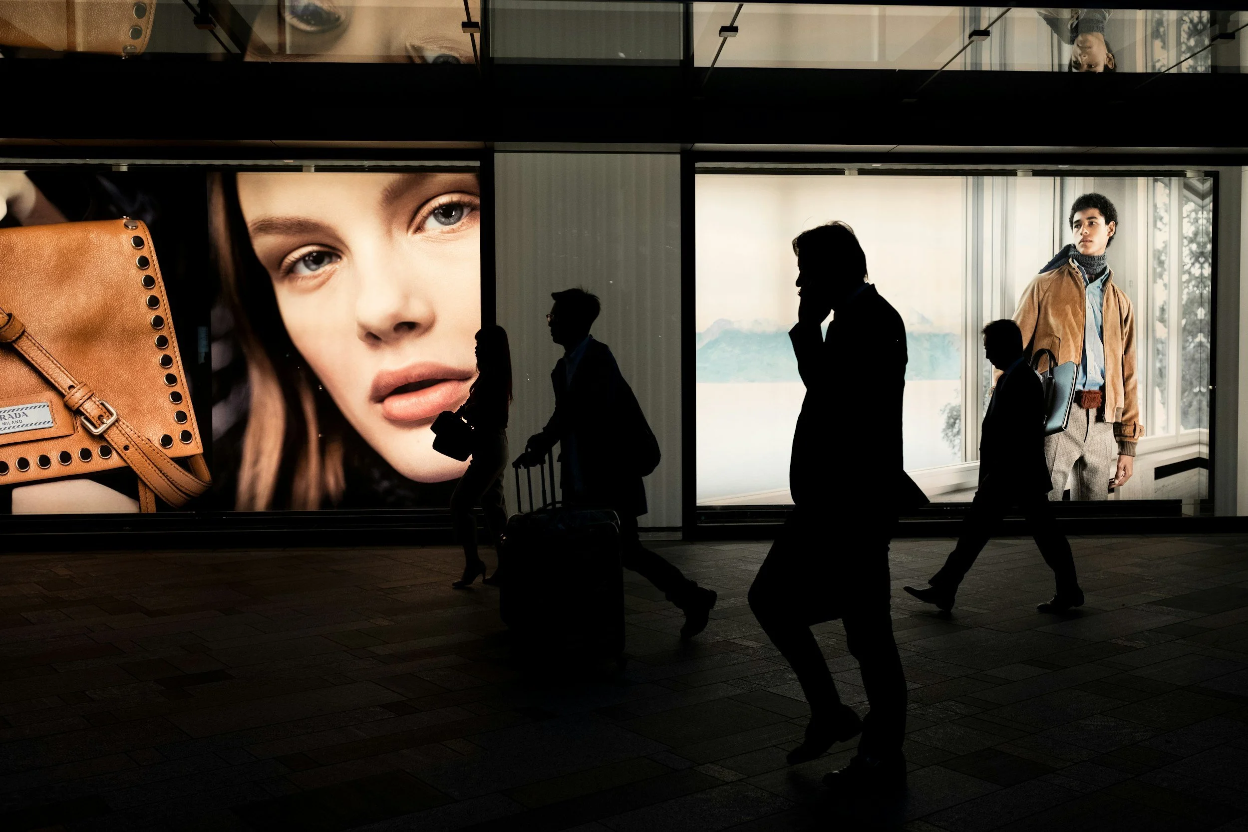 Silhouettes of five people walking past a large advertisement featuring a woman's face and a brown handbag at an airport or shopping mall.