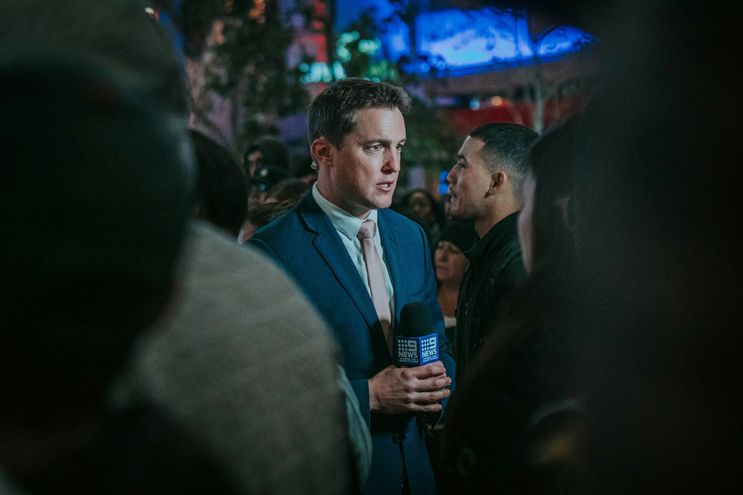 A man in a blue suit and white shirt holding a microphone with a blue logo is speaking to the media surrounded by crowd at night, with blurred colorful lights and trees in the background.