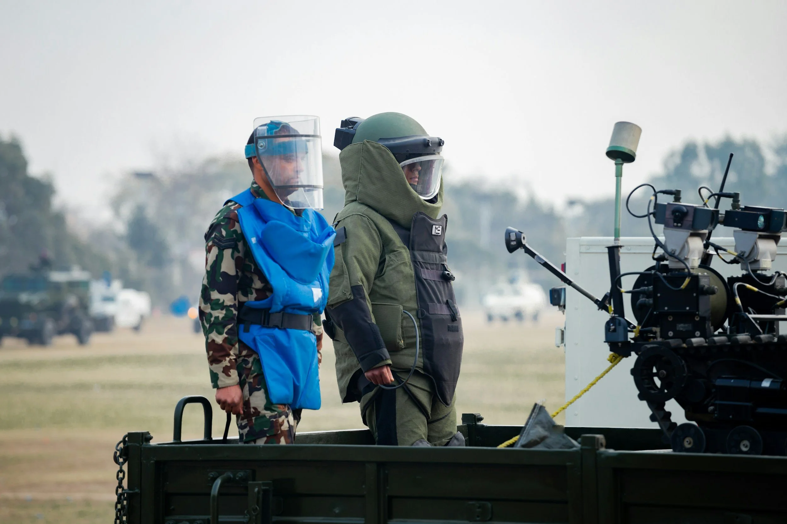 Two soldiers wearing protective gear and helmets stand beside a military vehicle at an outdoor field, with equipment and trees in the background.