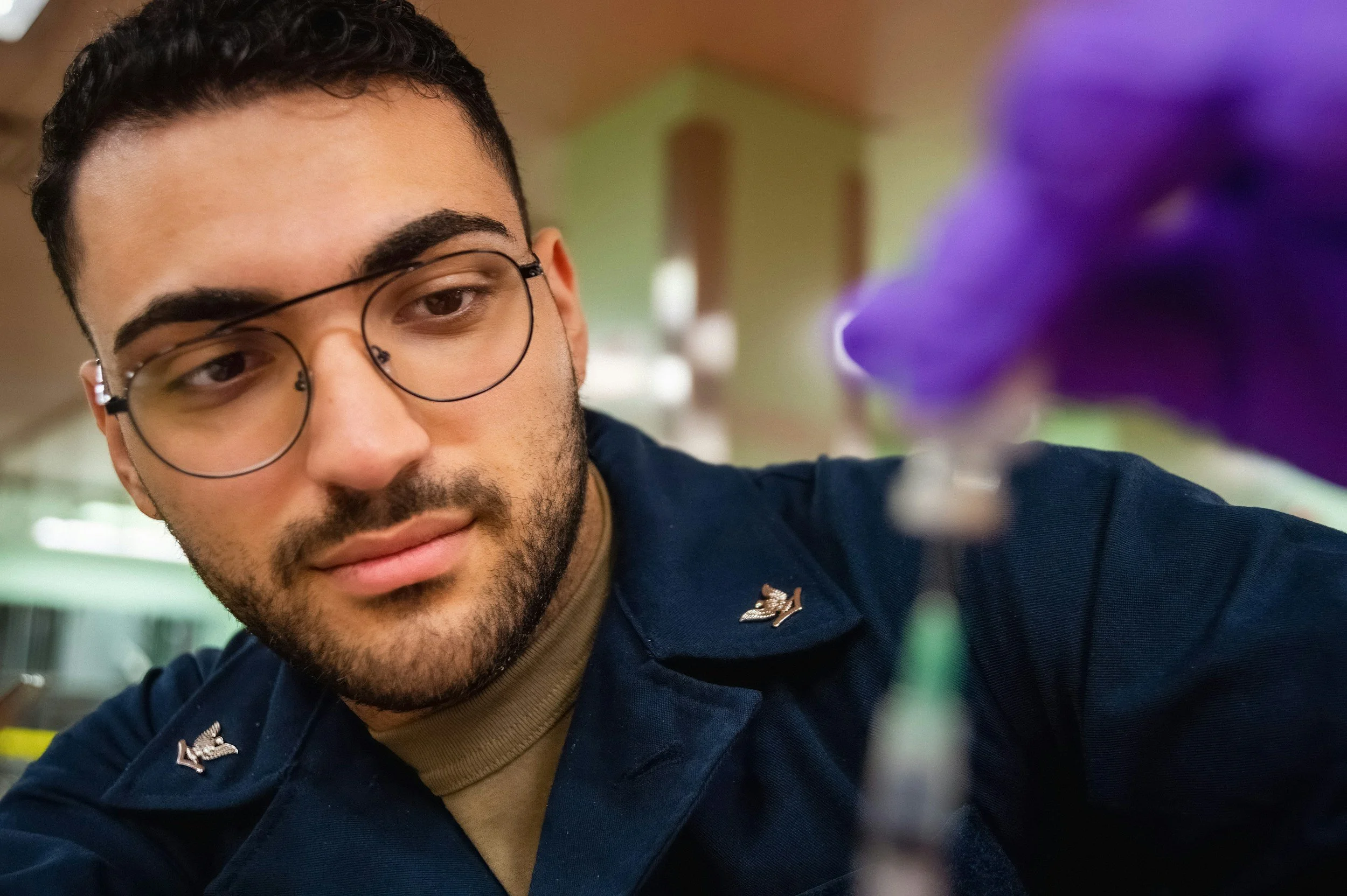 A close-up of a young man with glasses, short dark hair, a beard, and wearing a military-style uniform with a badge, looking slightly to his left, with a blurred purple flower in the foreground on the right.