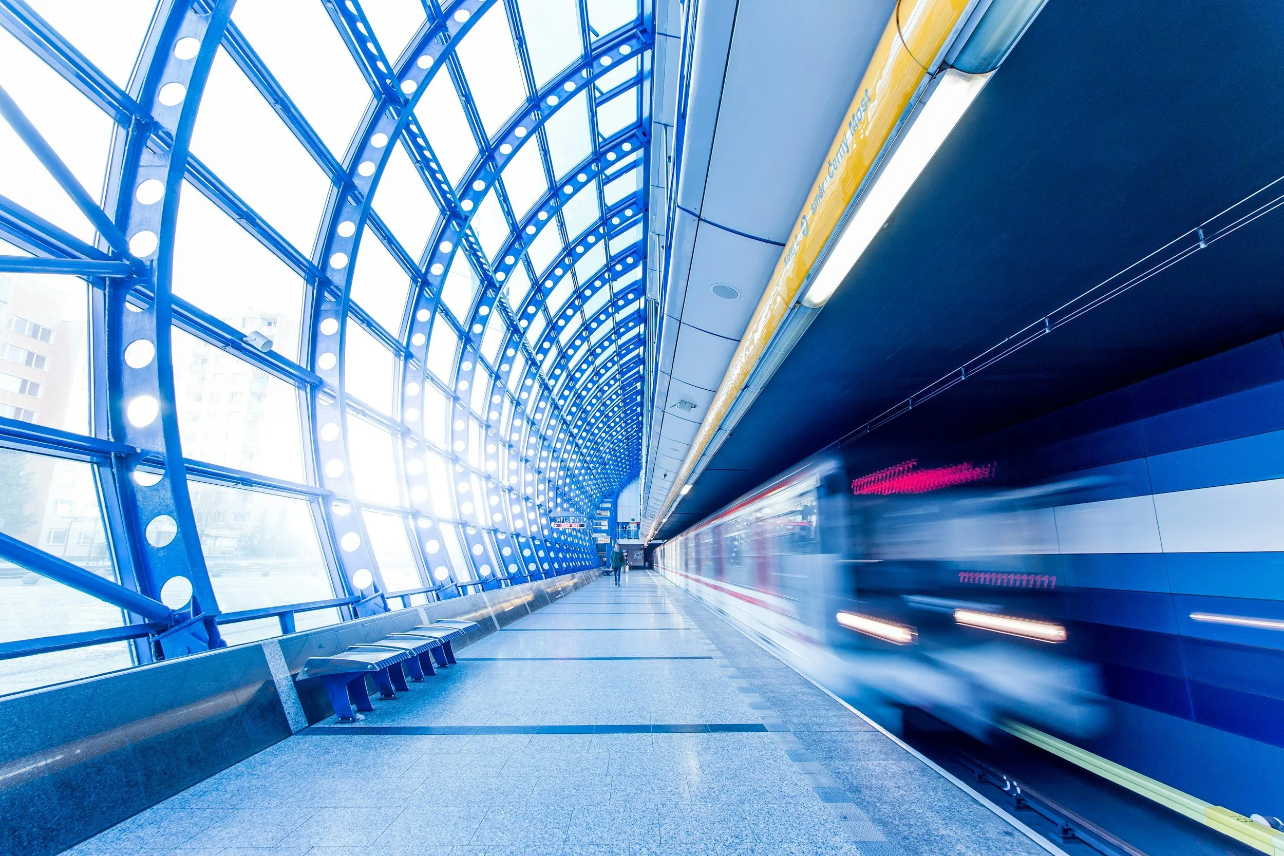 A motion-blurred train arriving at a modern subway station with blue architectural elements and a person walking in the distance.