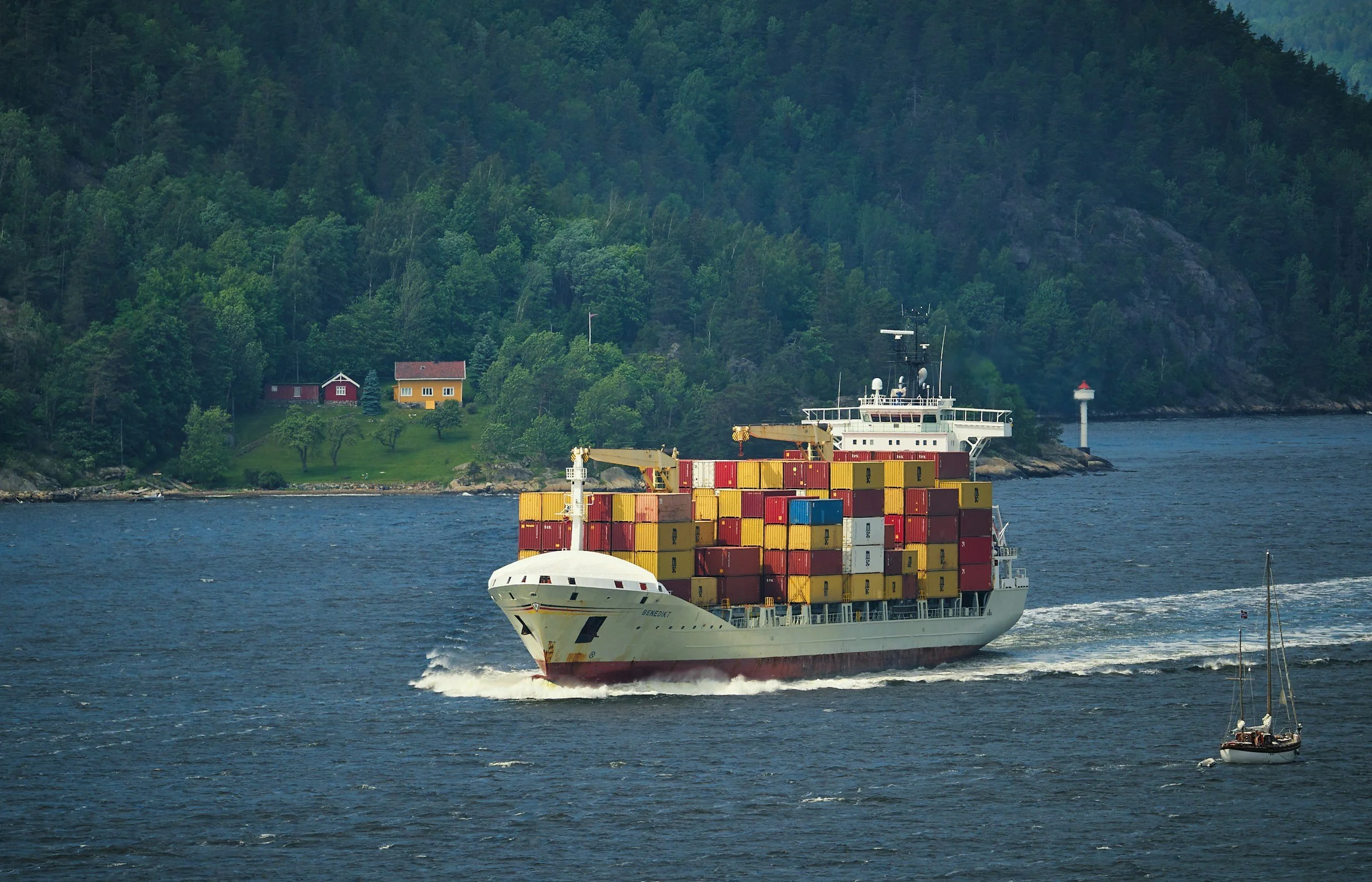 A large cargo ship carrying stacked shipping containers sailing in a body of water with green forested hills and small houses in the background.