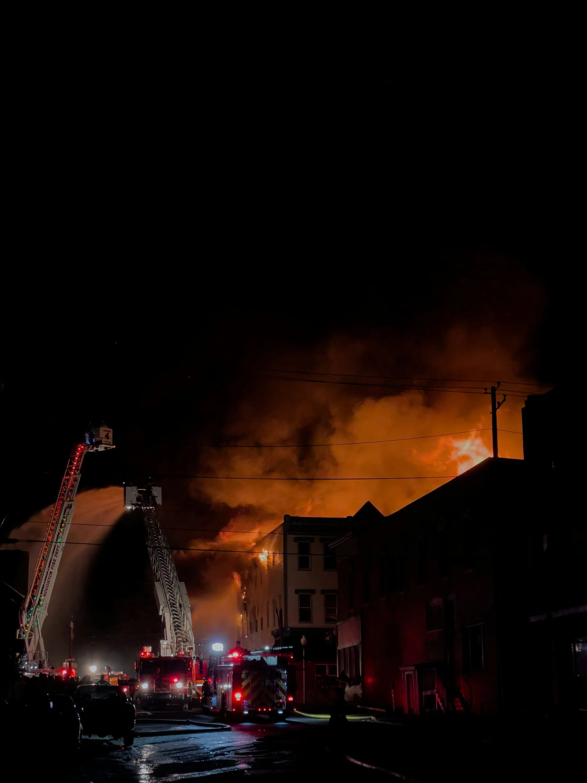 Firefighters respond to a major fire at night, with flames and thick smoke billowing from a building. Emergency vehicles and a ladder are visible, and residential buildings are in the foreground.