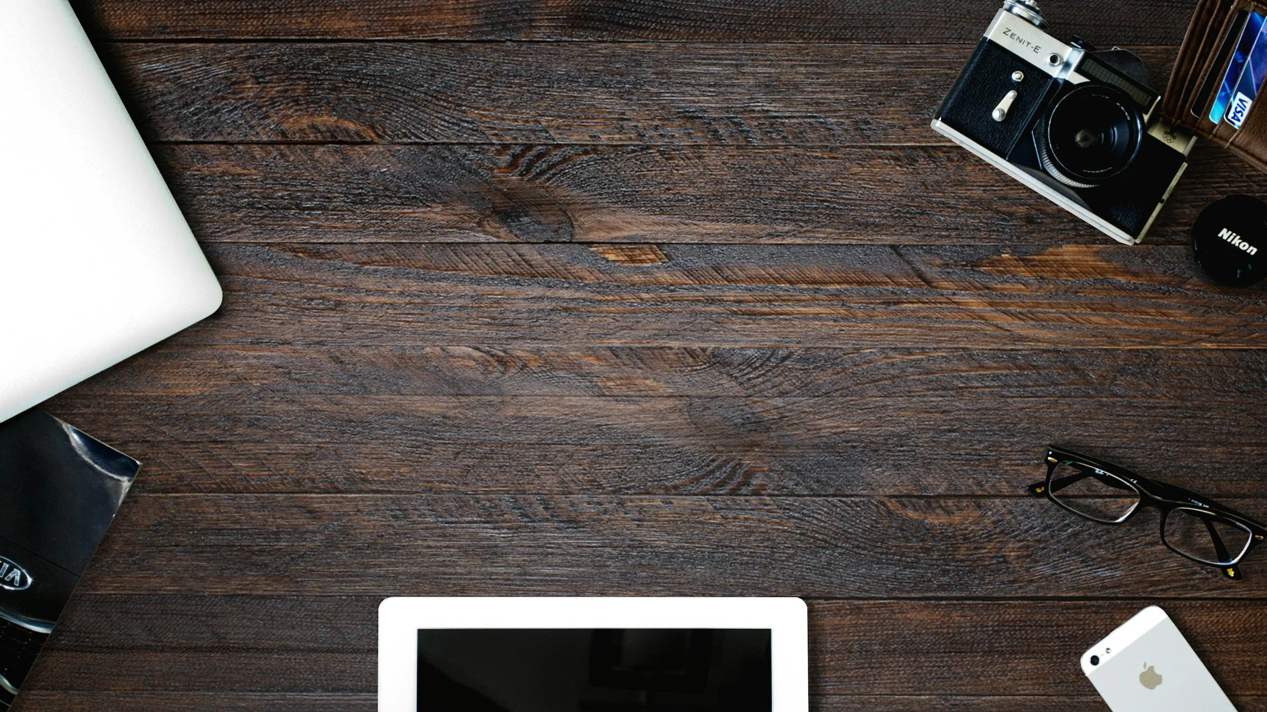 Top-down view of a dark wooden desk with electronic devices and accessories including a camera, glasses, phone, and a partial laptop.
