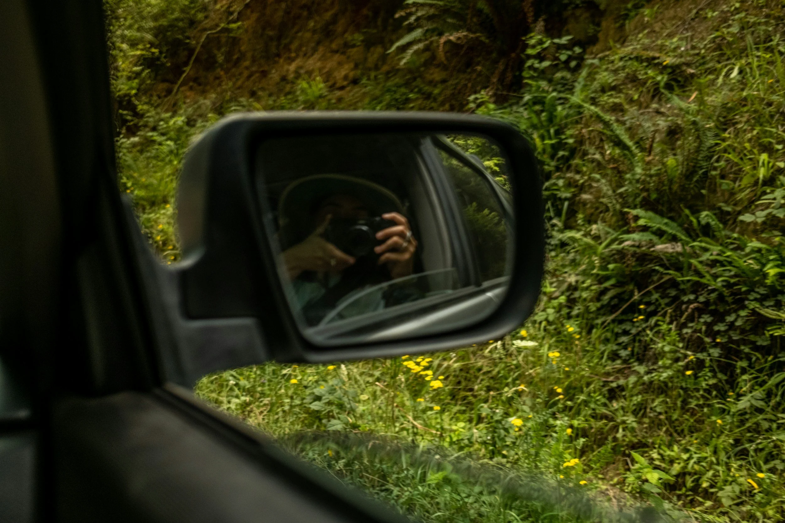 The image shows the view from inside a vehicle looking at a side mirror. In the mirror, a person is taking a photo with a camera. Outside, there is green foliage and a dirt path or road.