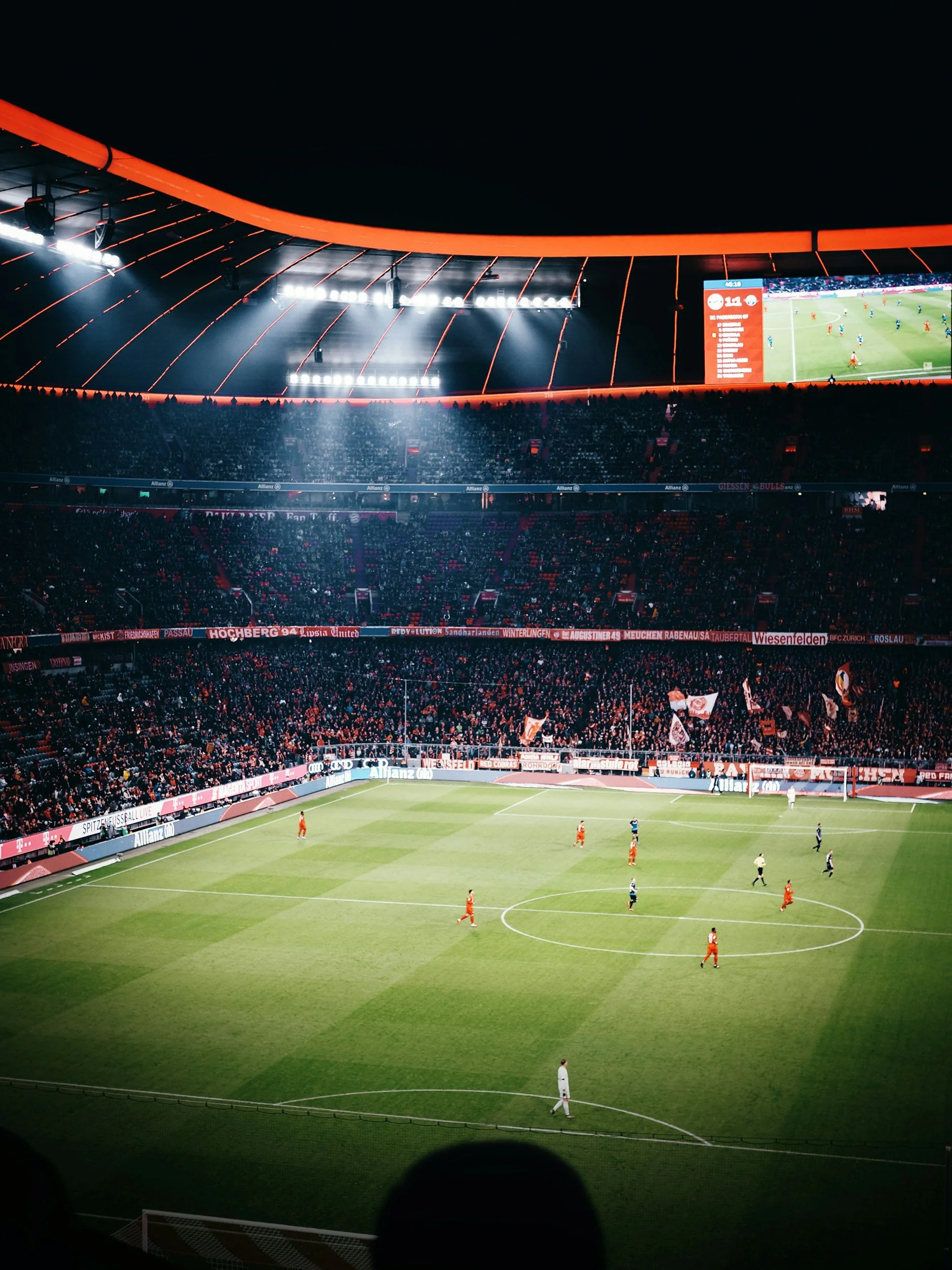 A soccer stadium filled with fans, players on the field, bright stadium lights, and large video screens displaying the ongoing match at night.