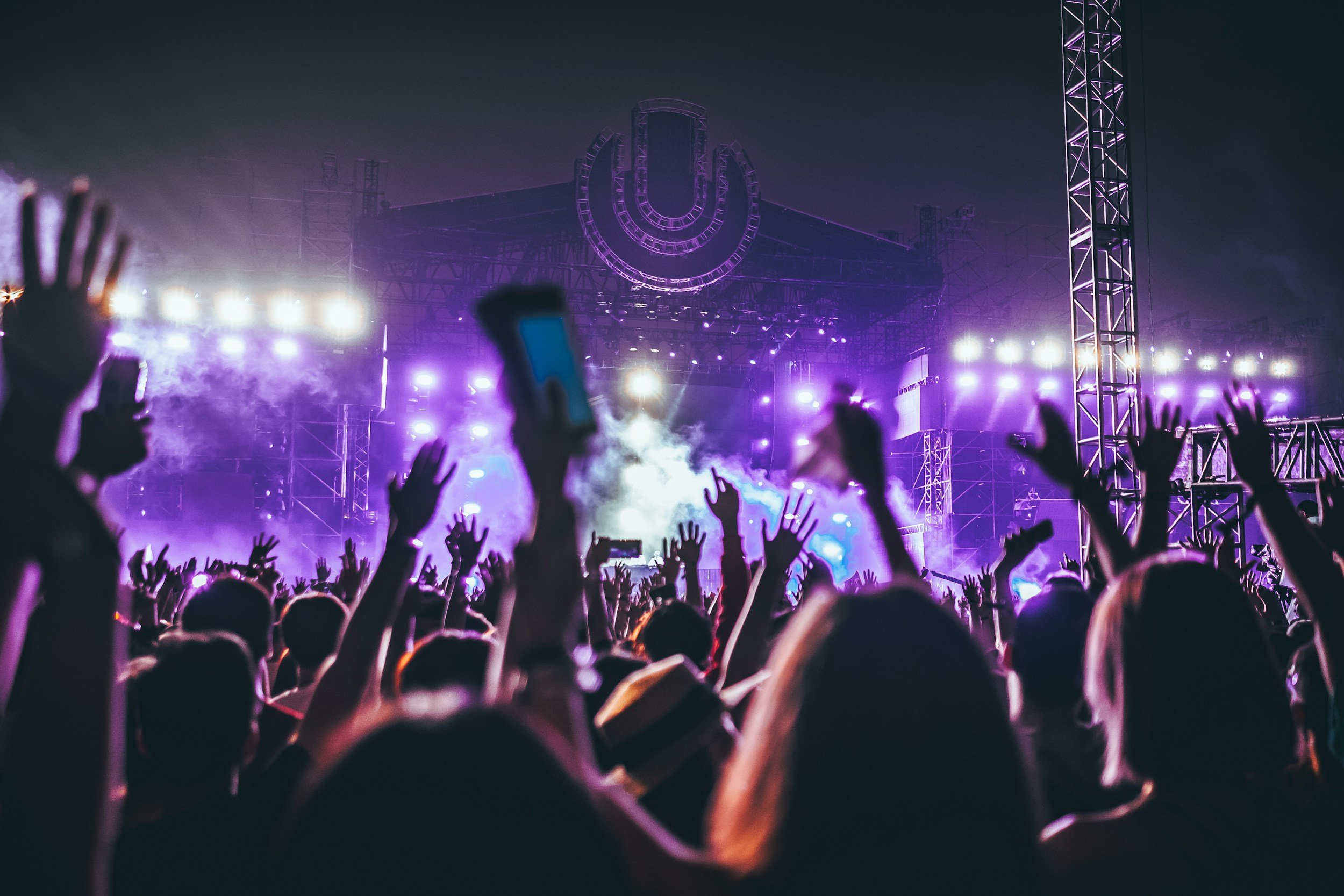 A crowd at a concert or music festival with hands raised, purple stage lighting, and a large illuminated symbol in the background.