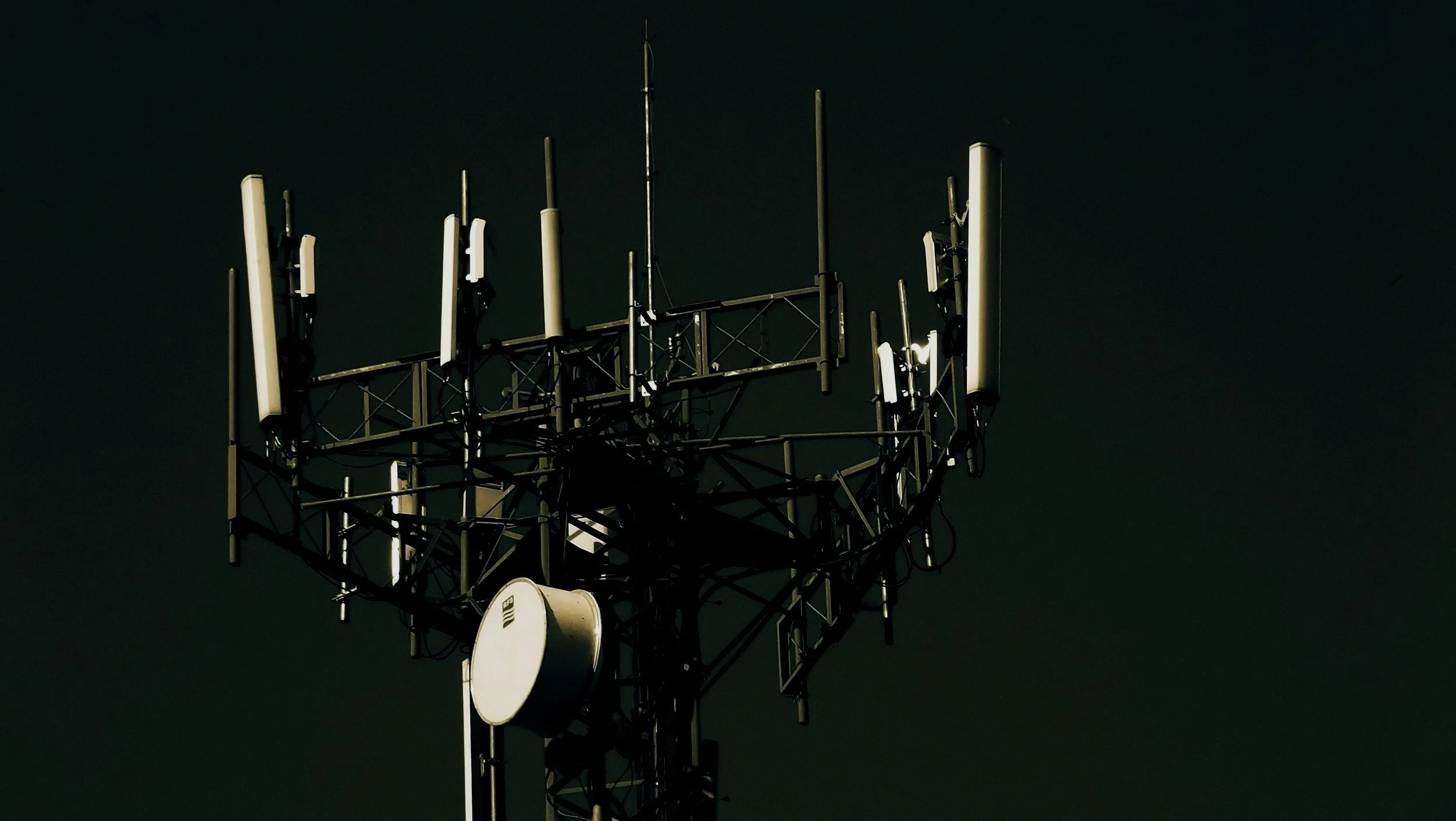 A telecommunications tower with multiple antennas and dishes against a dark sky.