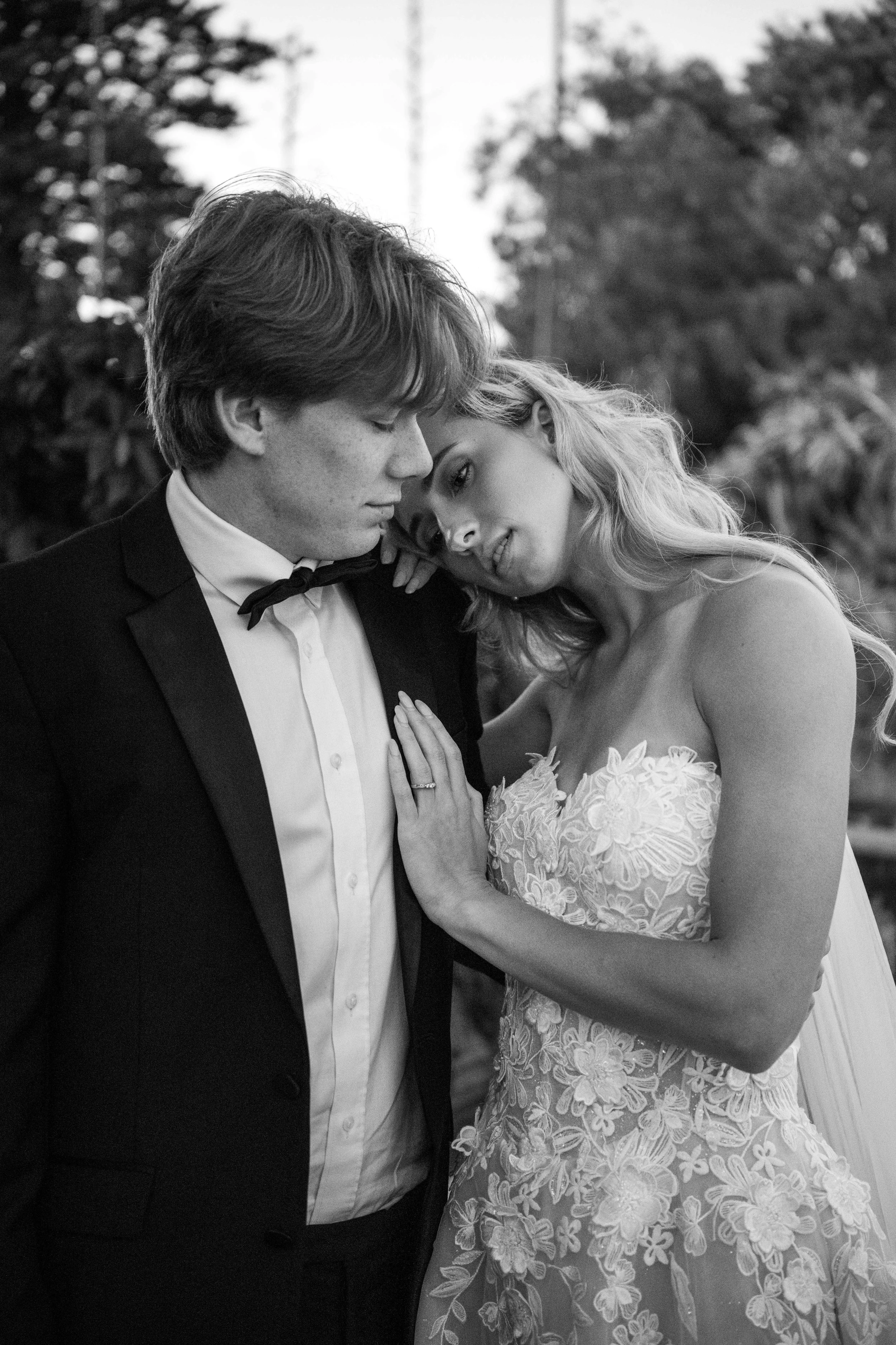 A black-and-white photograph of a bride and groom sharing an emotional moment. The bride, wearing a lace wedding gown, gently touches the groom's chest, while the groom, dressed in a tuxedo with a bow tie, leans his forehead against hers.