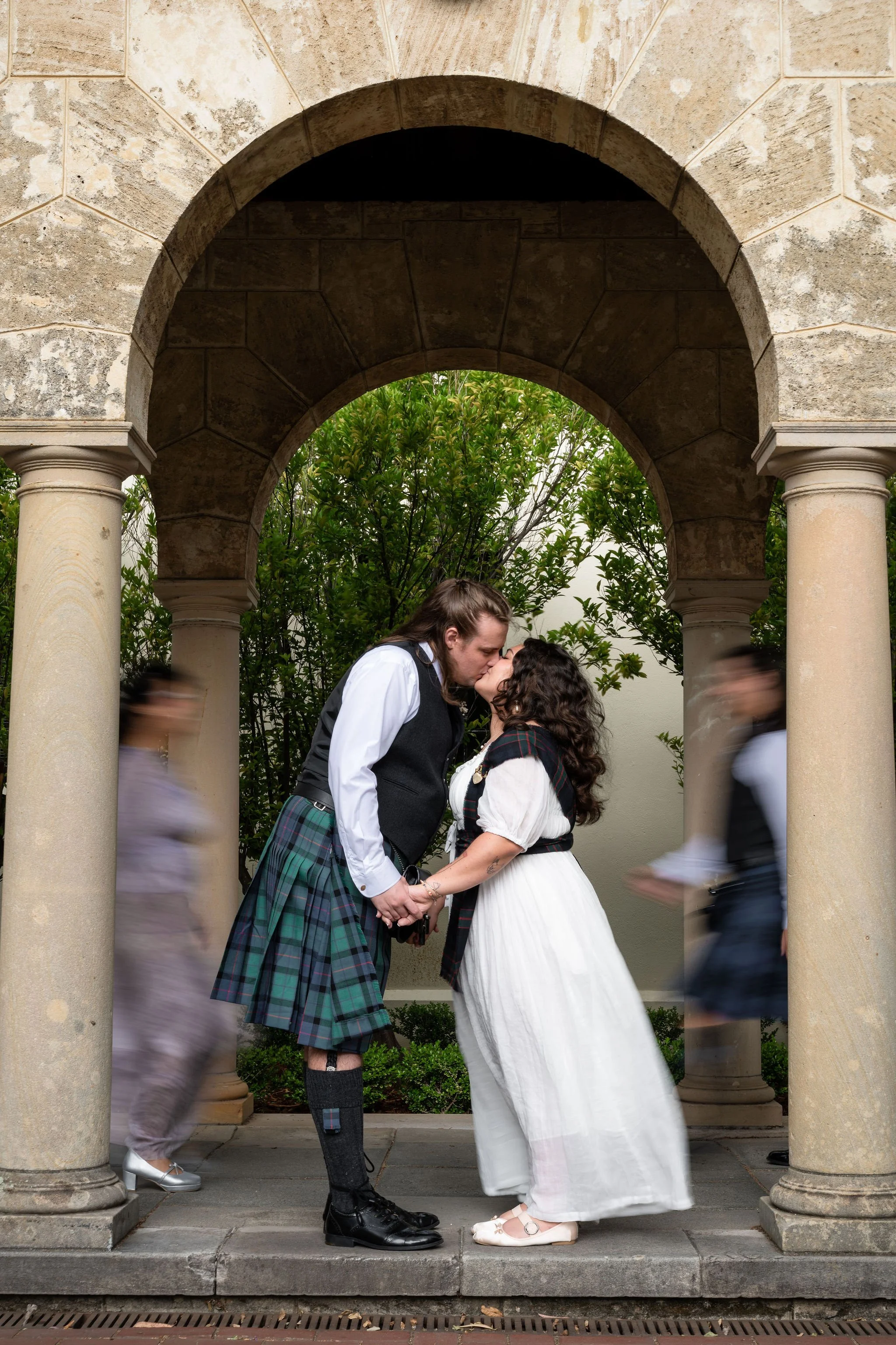 A couple dressed in Scottish attire sharing a kiss under an archway, with blurred pedestrians walking by.