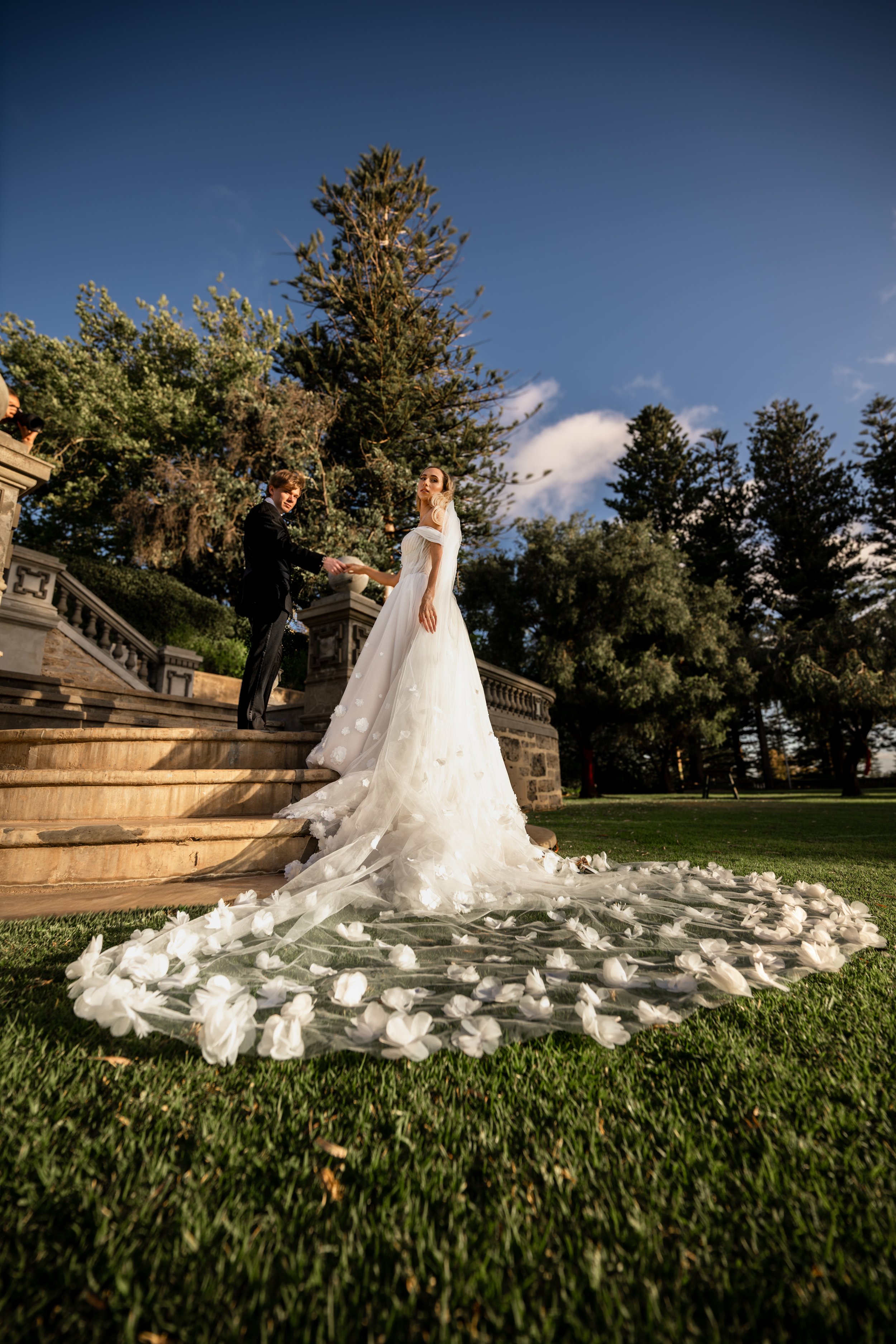 Bride and groom exchanging vows on outdoor stairs with bride's floral train on grass and wedding arch in background, trees, and blue sky.