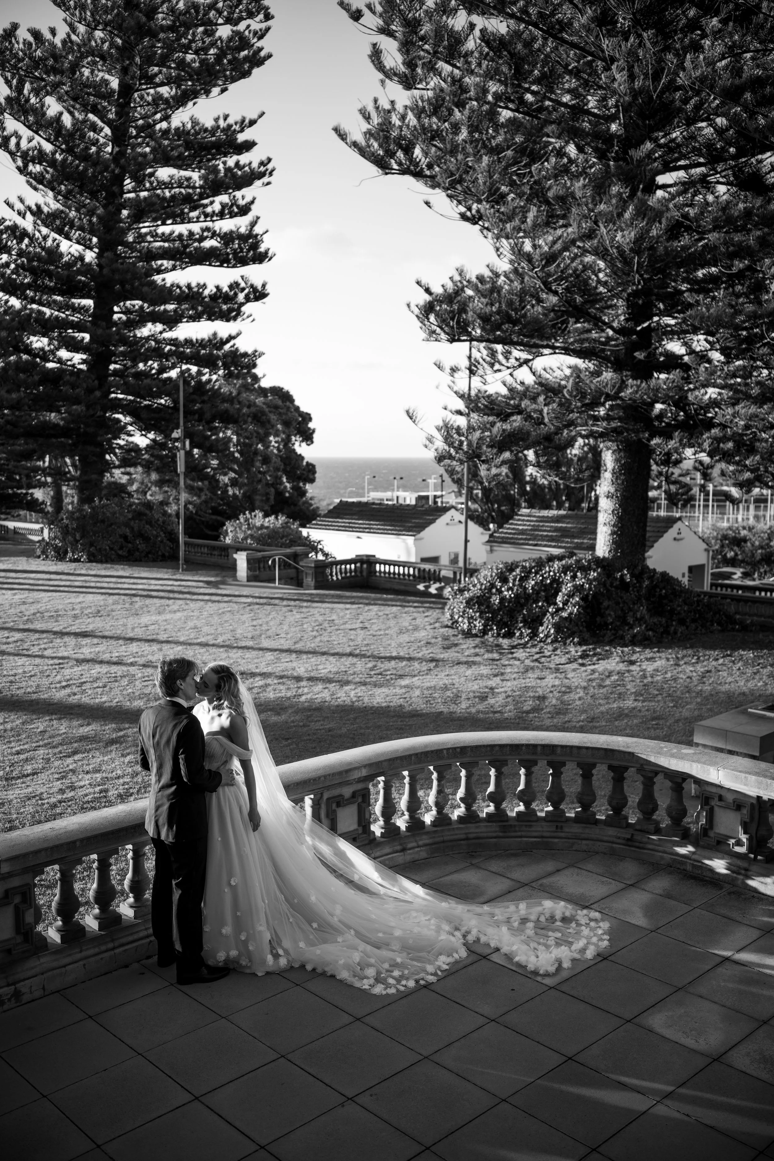 A black and white photo of a bride and groom kissing on a terrace with a scenic outdoor background of trees, shrubs, and rooftops. The bride is wearing a long flowing wedding dress with a train, and the groom is in a suit.