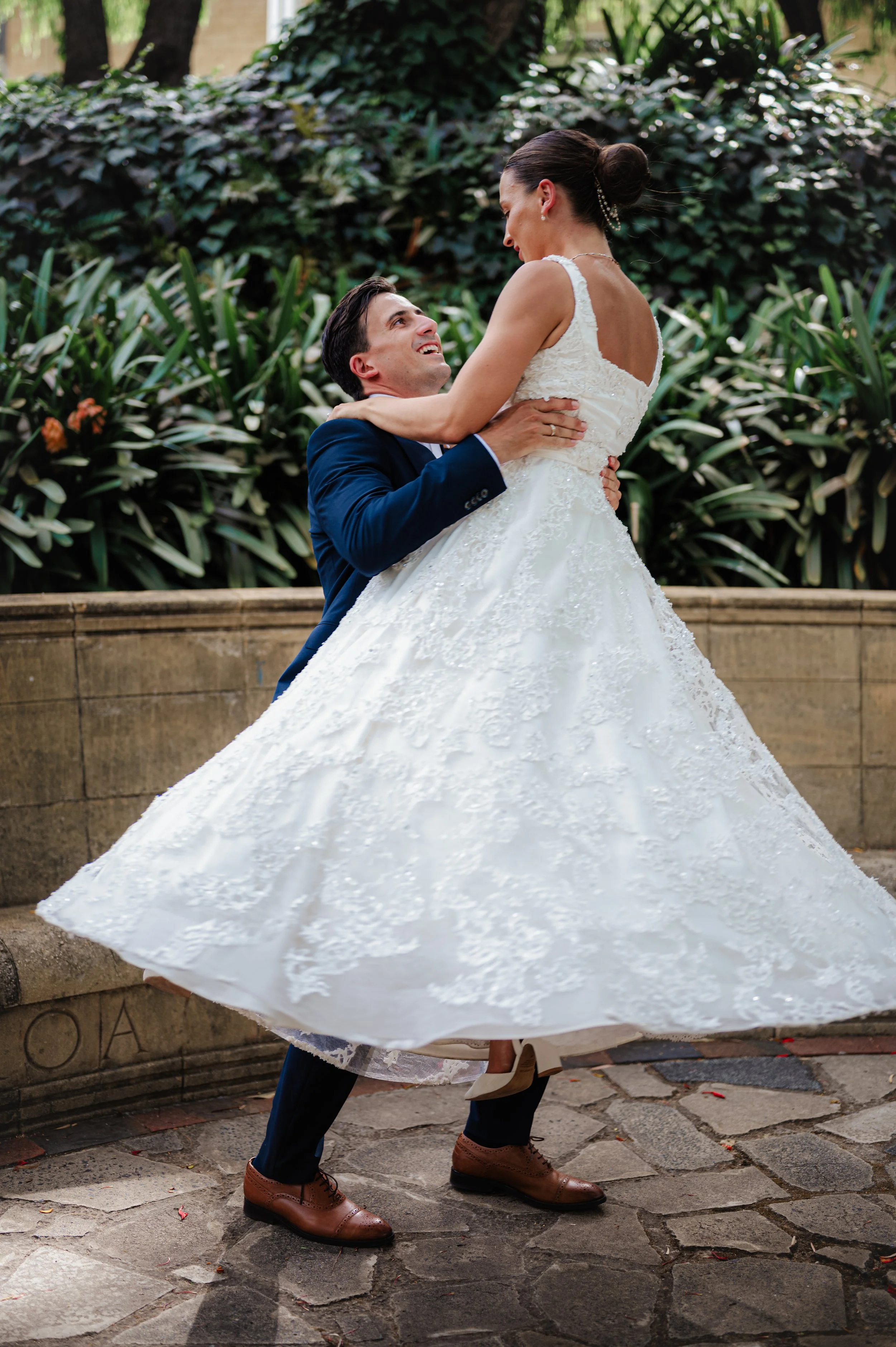 A bride and groom celebrate outdoors, with the groom lifting the bride in his arms as she holds him around his neck, both looking at each other happily.