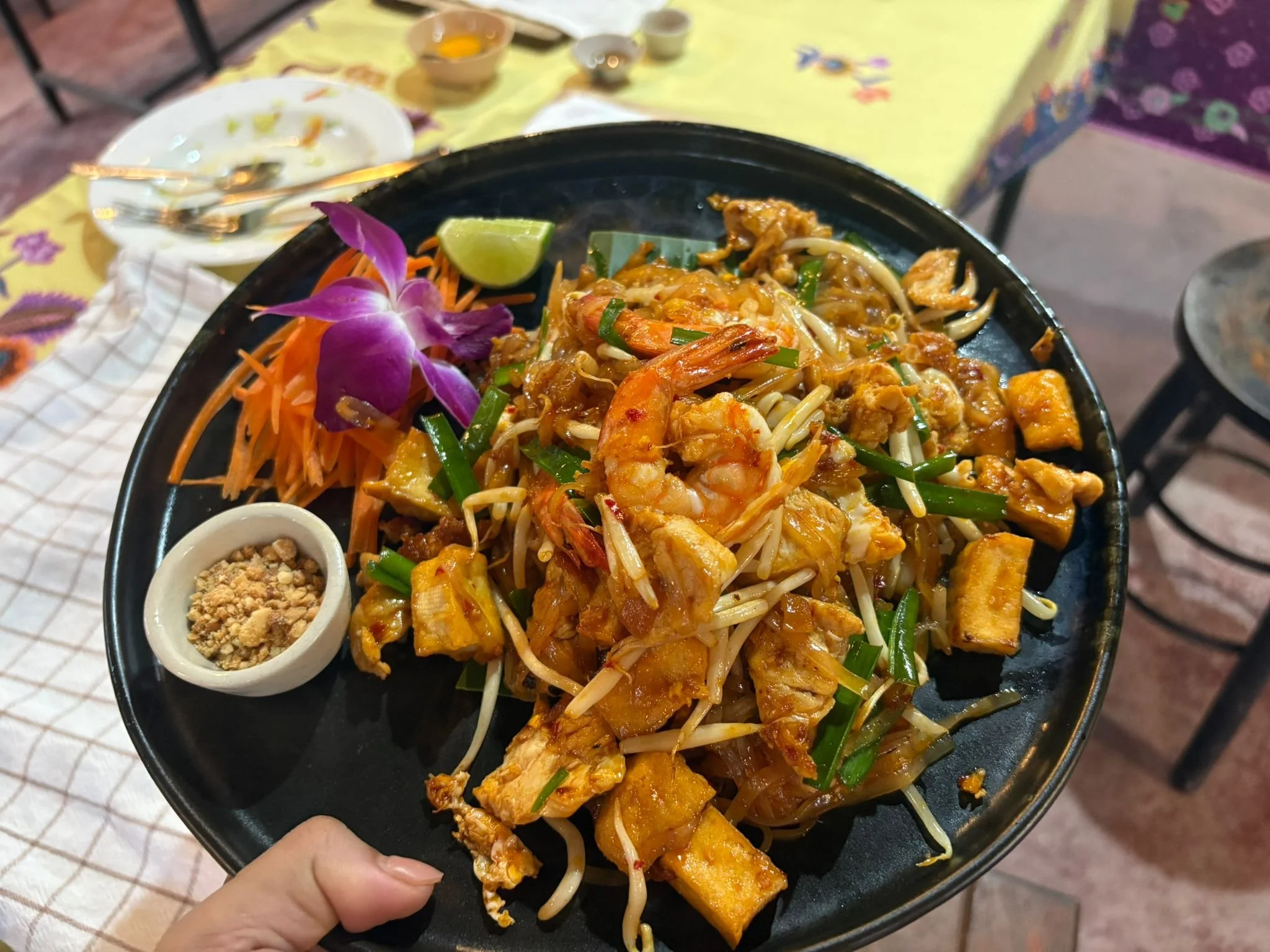 Plate of stir-fried noodles with shrimp, chicken, bean sprouts, green onions, and a lime wedge, garnished with a purple orchid and shredded carrots, with condiments and utensils on a table.
