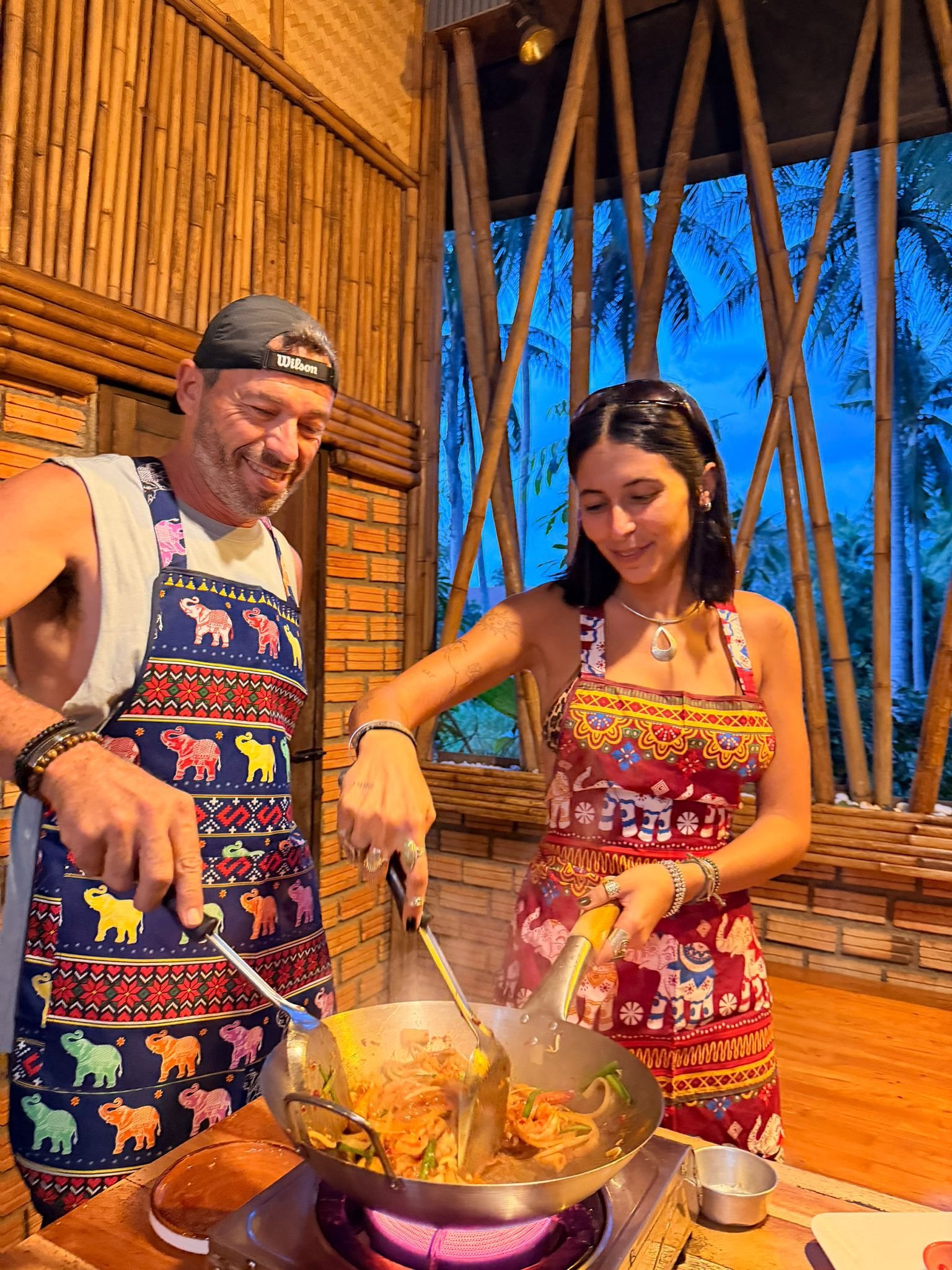 Two people cooking together in a bamboo-themed kitchen, stirring a large pan of food on a stove, with a blue sky visible through the open window behind them.