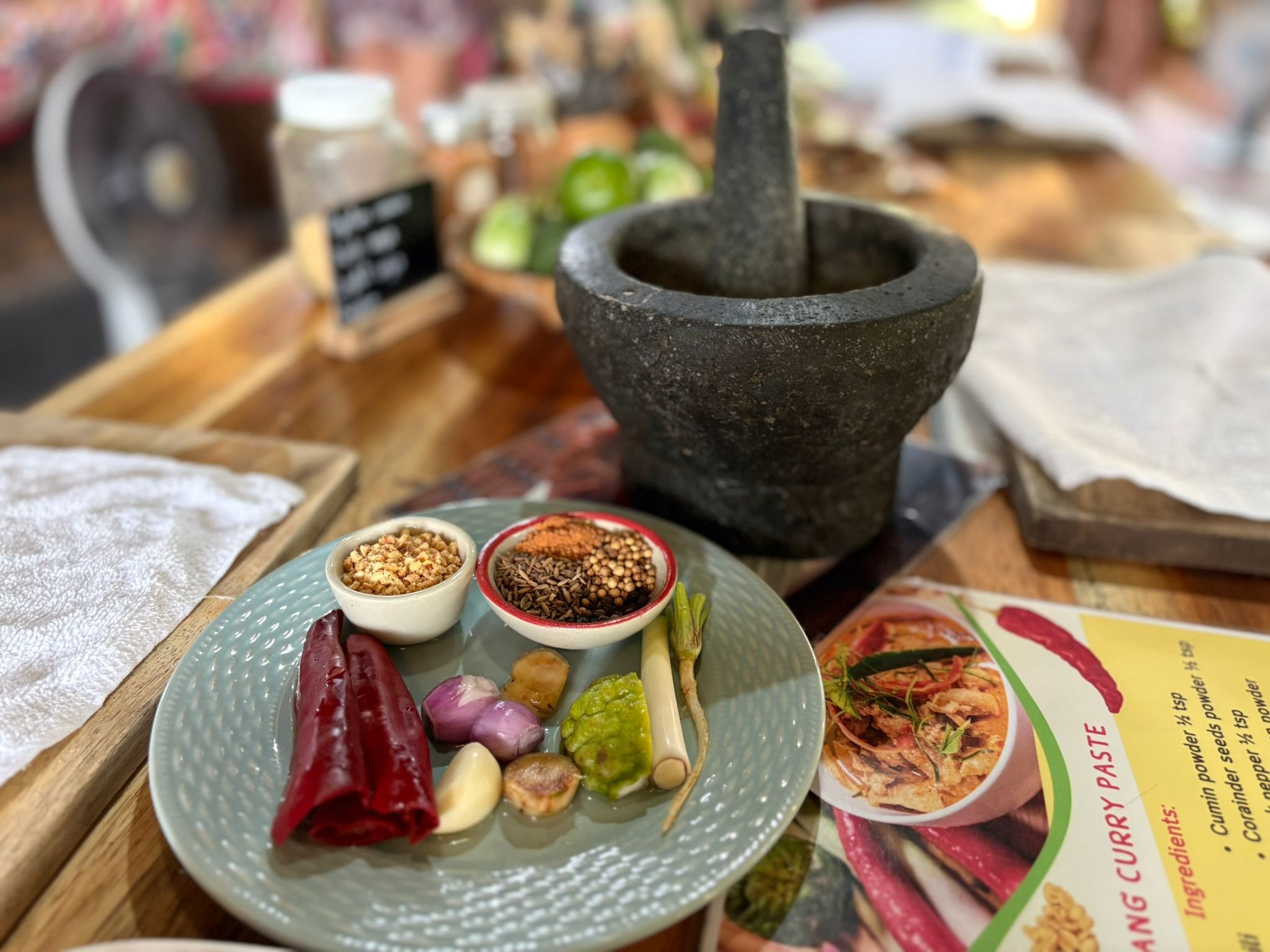 A plate with various spices, garlic, shallots, and chili peppers, with a mortar and pestle nearby, set on a wooden table at a restaurant or market.