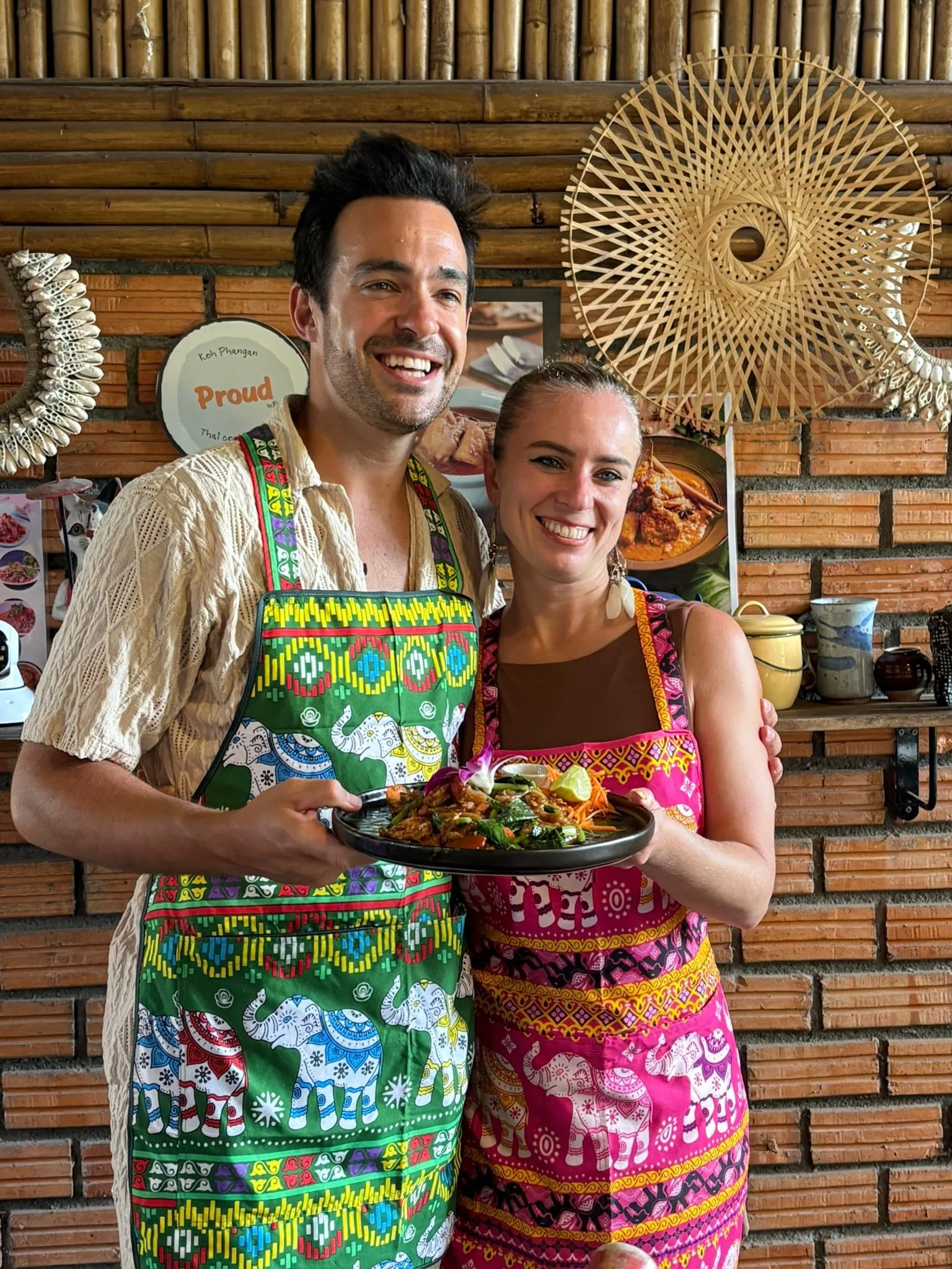A smiling man and woman wearing colorful aprons with elephant patterns, holding a plate of food in a rustic restaurant with wooden walls and decorative bamboo art.