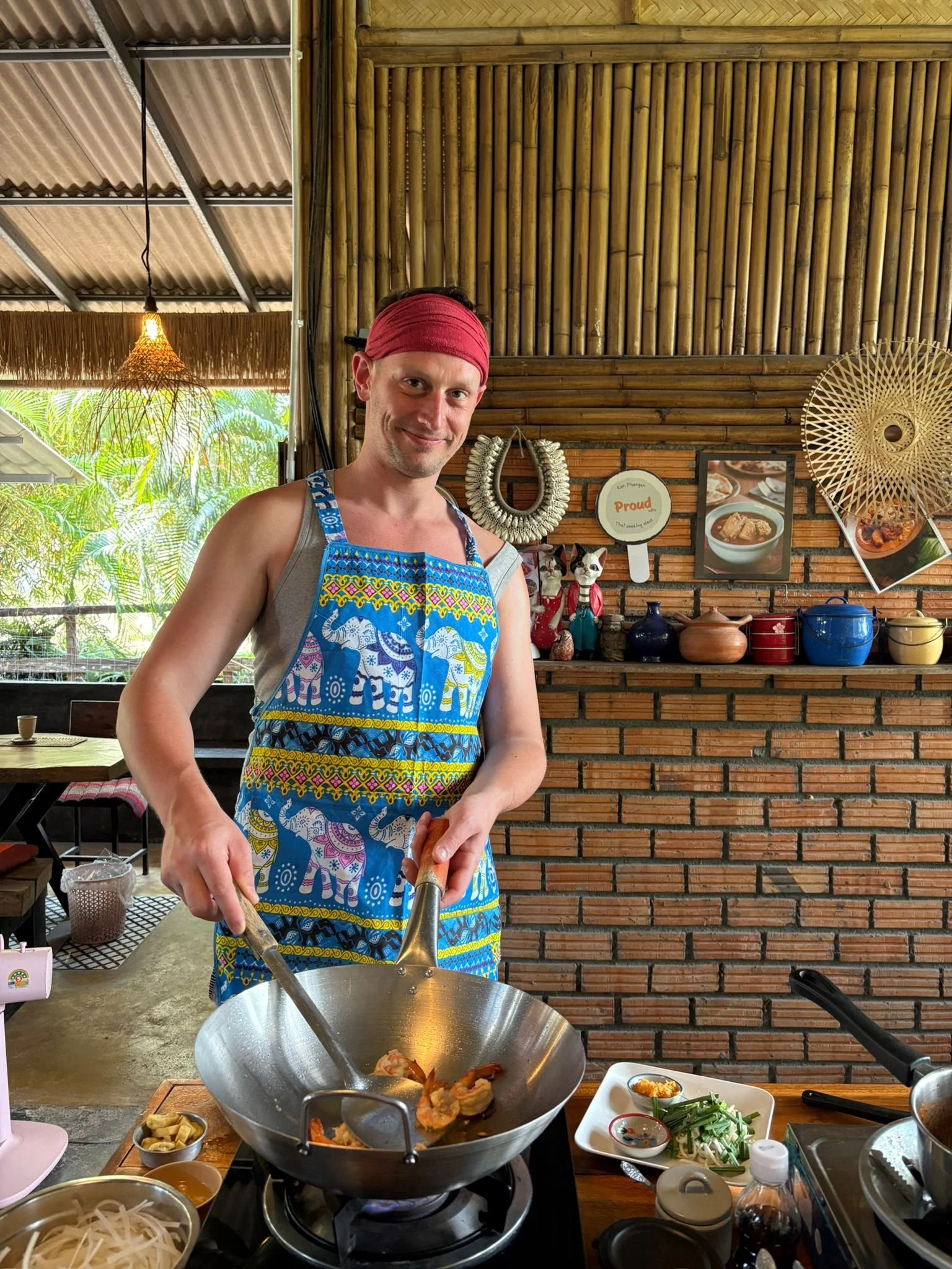 A man wearing a colorful apron with elephants, a gray tank top, and a red headband cooking in a kitchen with bamboo and brick decor.