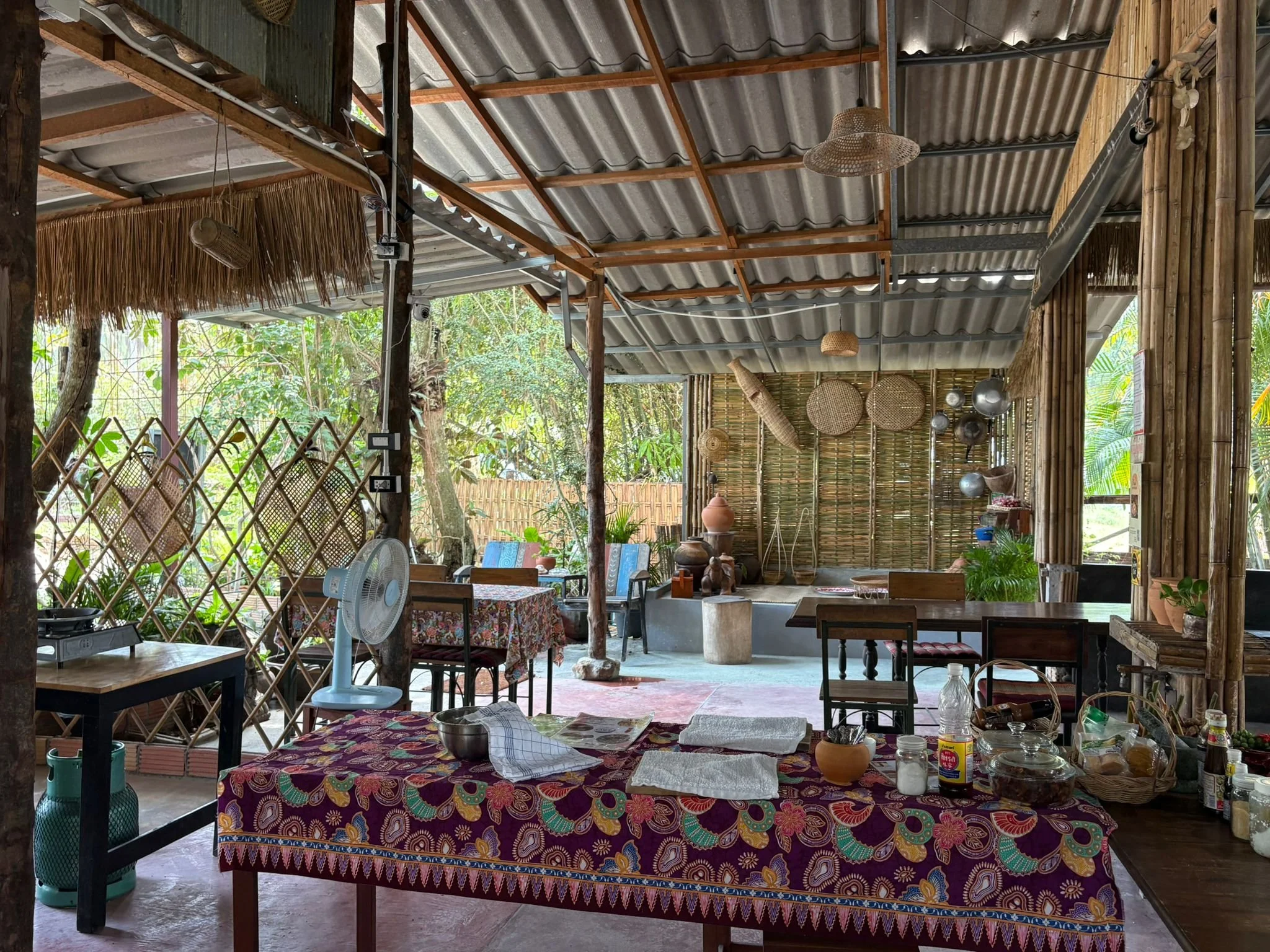 Interior of a rustic, open-air space with bamboo and thatch decor, a table with a colorful tablecloth, kitchen items, and outdoor greenery visible through the sides.