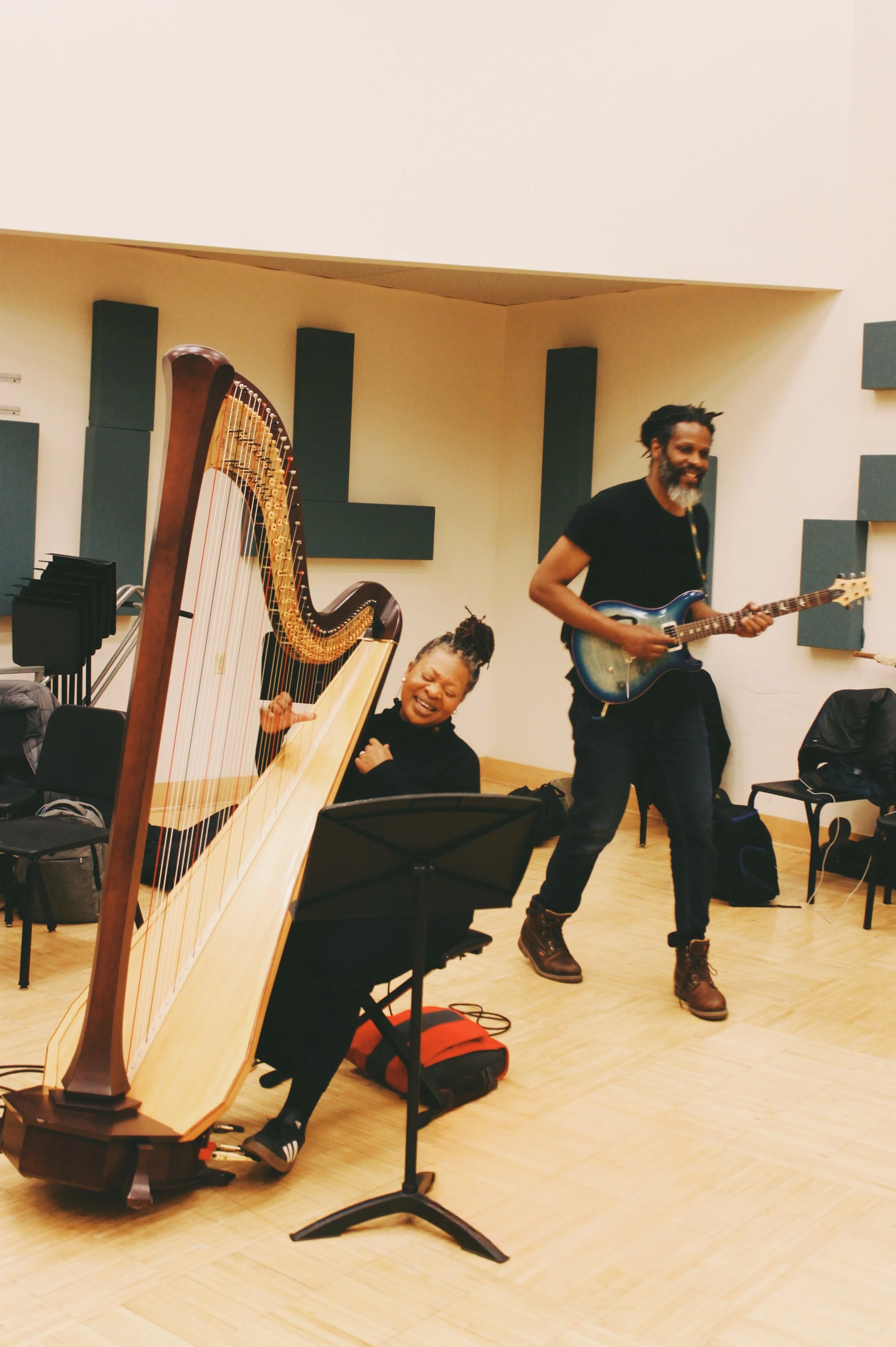 Two musicians performing in a room with a harp and an electric guitar. The woman is playing the harp and appears to be singing or enjoying the music with her eyes closed, while the man is standing and playing the guitar, smiling.