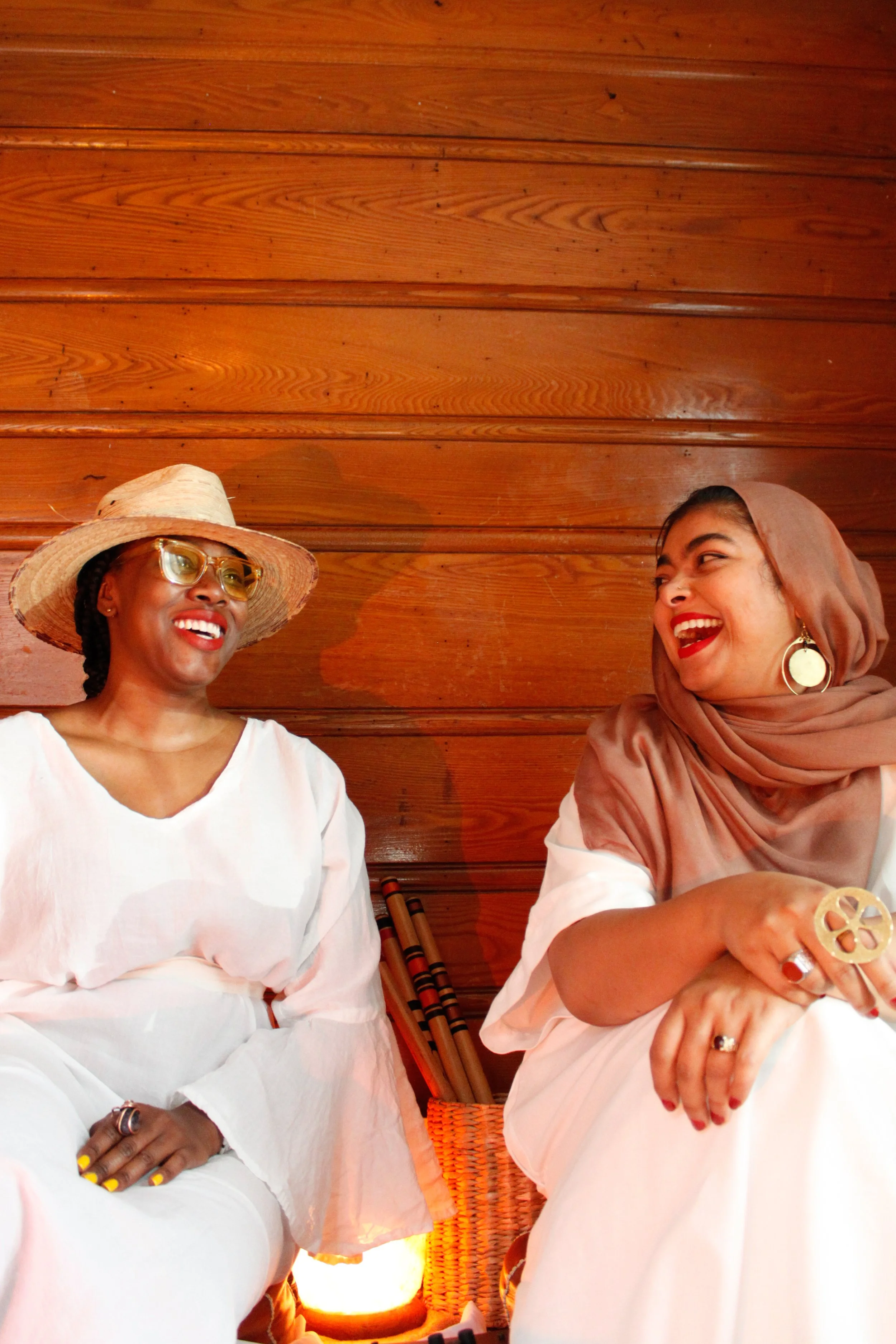 Two women sitting next to each other in front of a wooden wall, smiling and laughing, engaged in conversation.