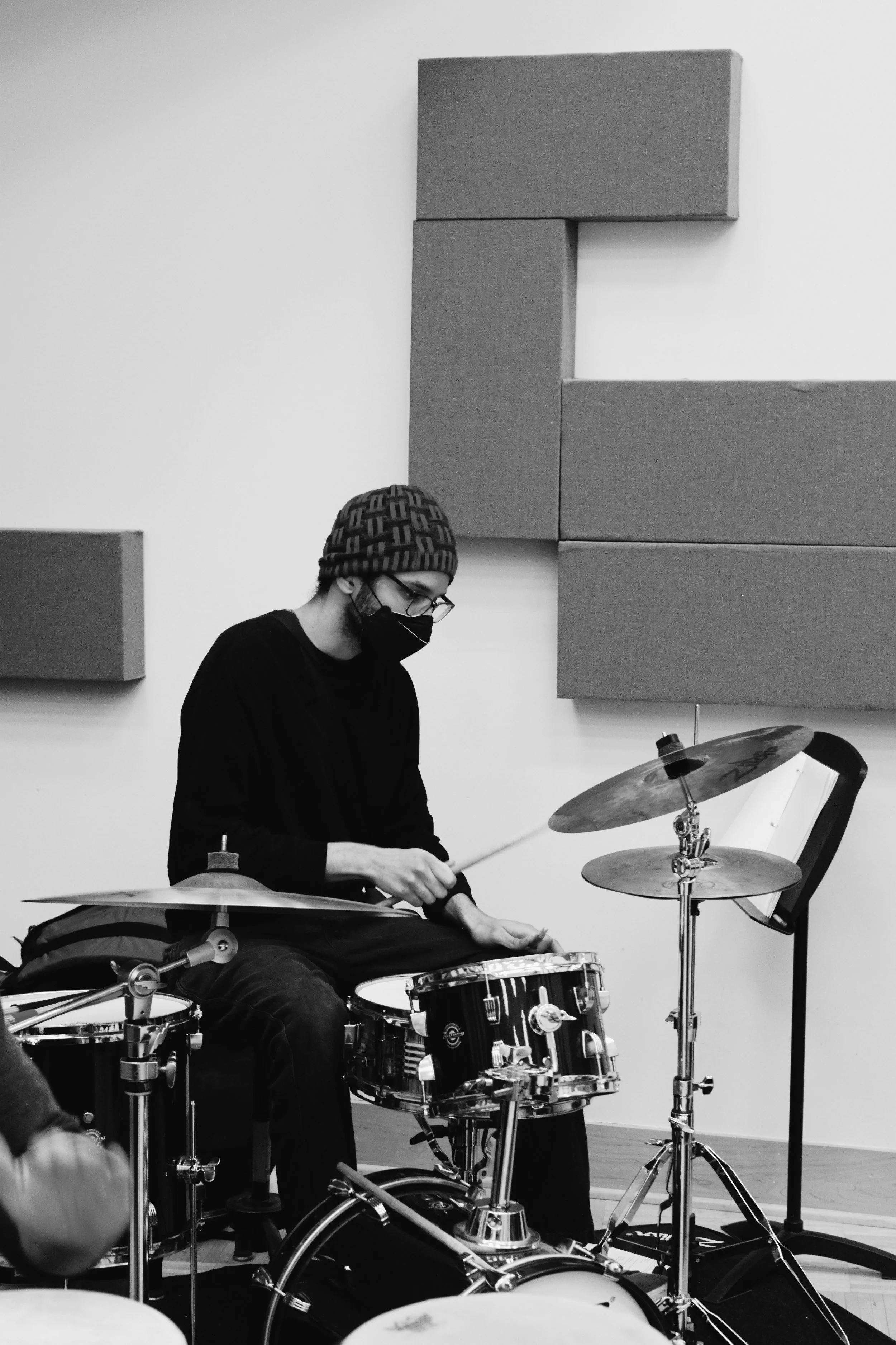 A person in a cap and face mask playing drums in a music studio with acoustic panels on the wall.
