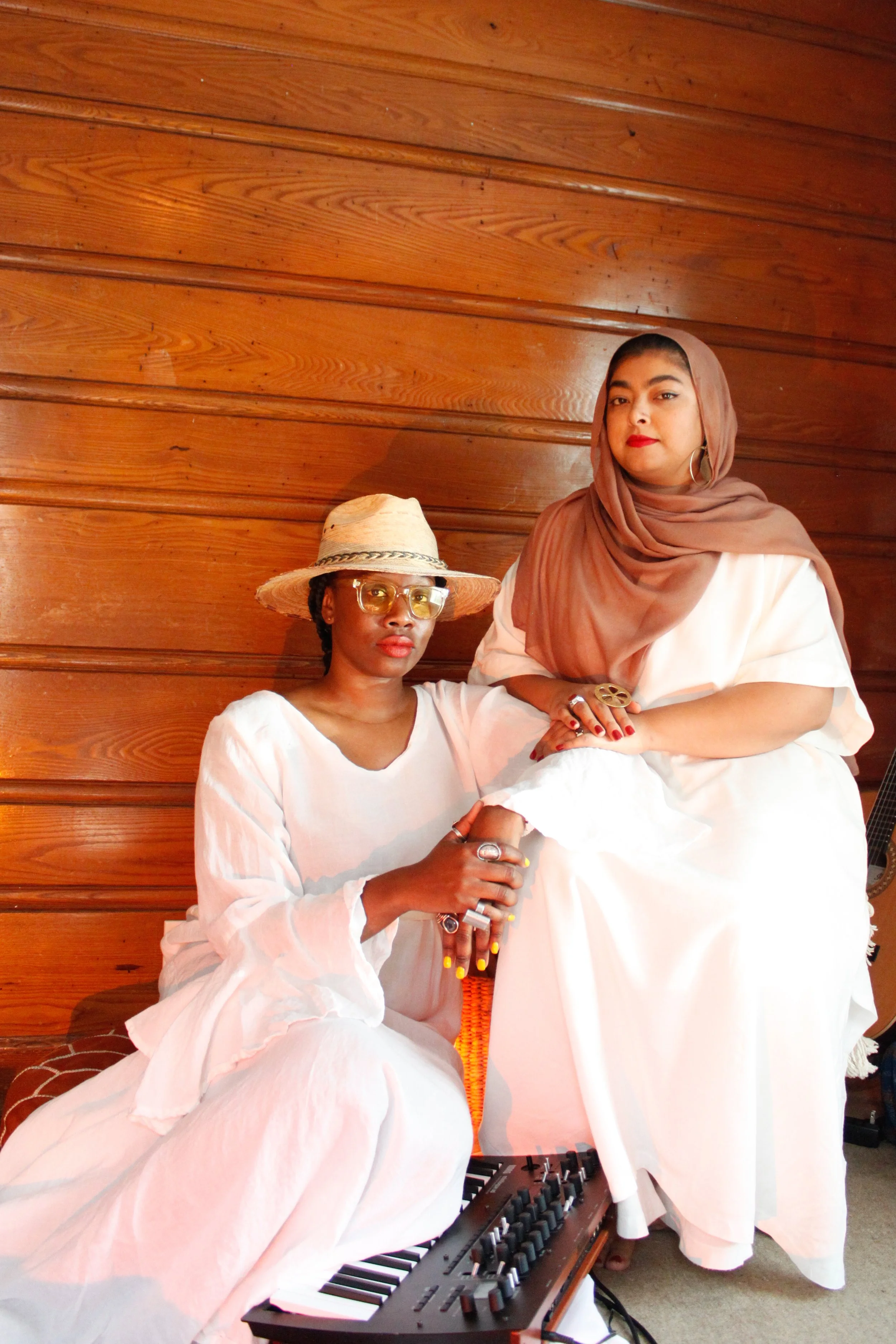 Two women wearing white dresses, one seated with a digital keyboard, and one standing, both against a wooden wall. The seated woman wears a wide-brimmed hat and glasses, while the standing woman has a brown headscarf and hoop earrings.