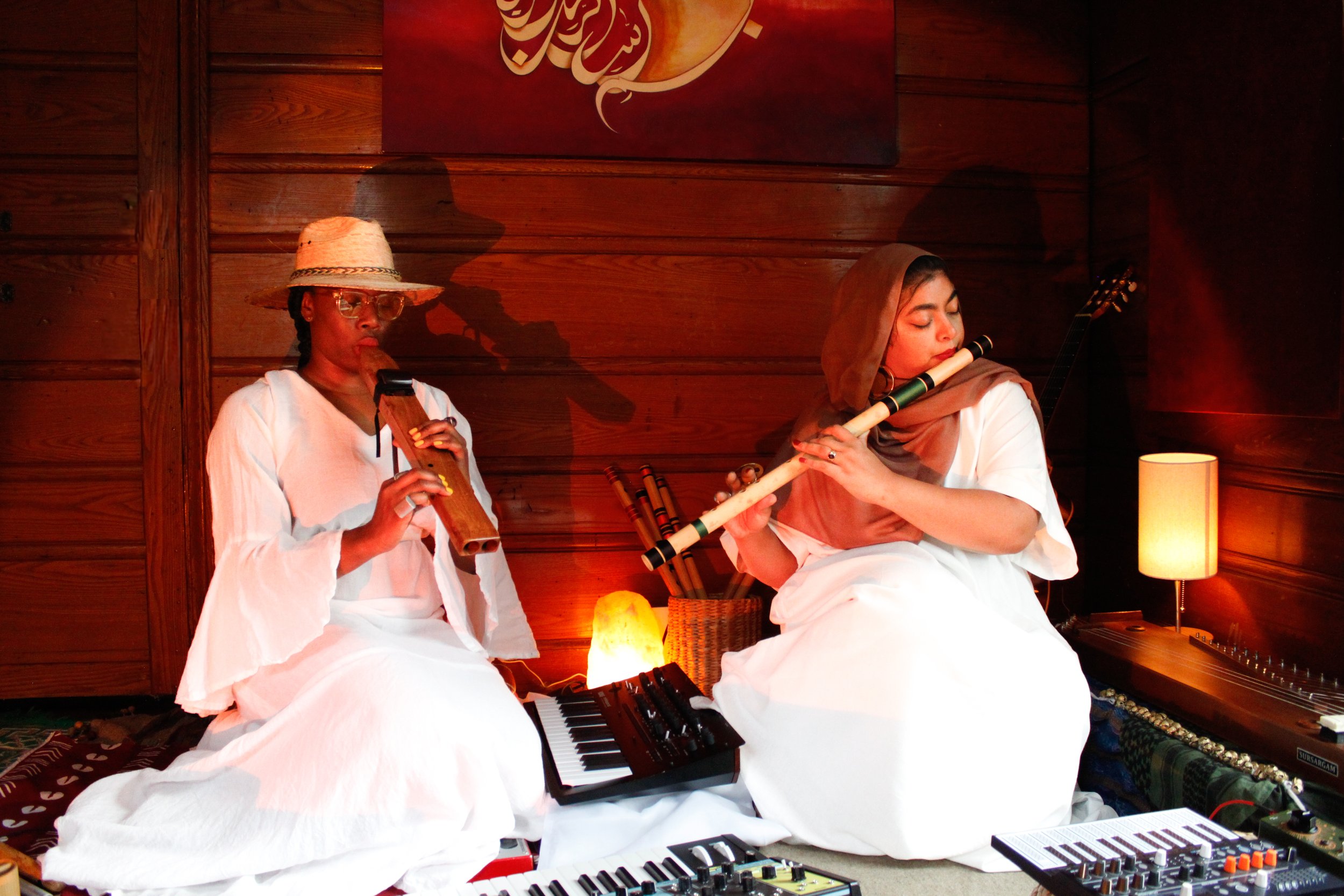 Two women sitting on the floor in a cozy wooden room, playing musical instruments: one with a bamboo flute and the other with a recorder. They are dressed in white flowing dresses, with one wearing a straw hat and glasses, and the other wearing a hea