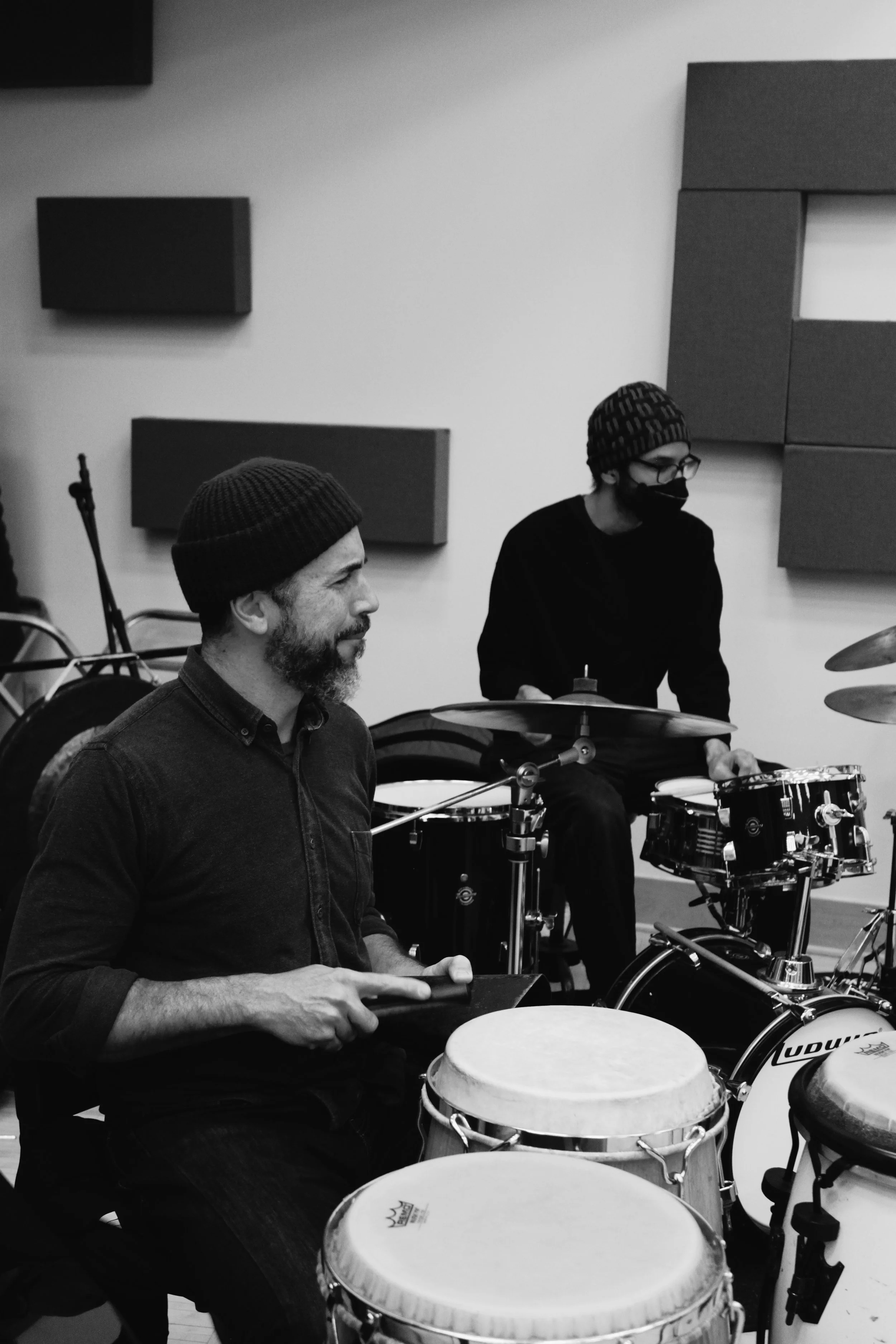 Two men playing percussion instruments in a music studio, one with a beard and a beanie, and the other wearing glasses, a mask, and a beanie.