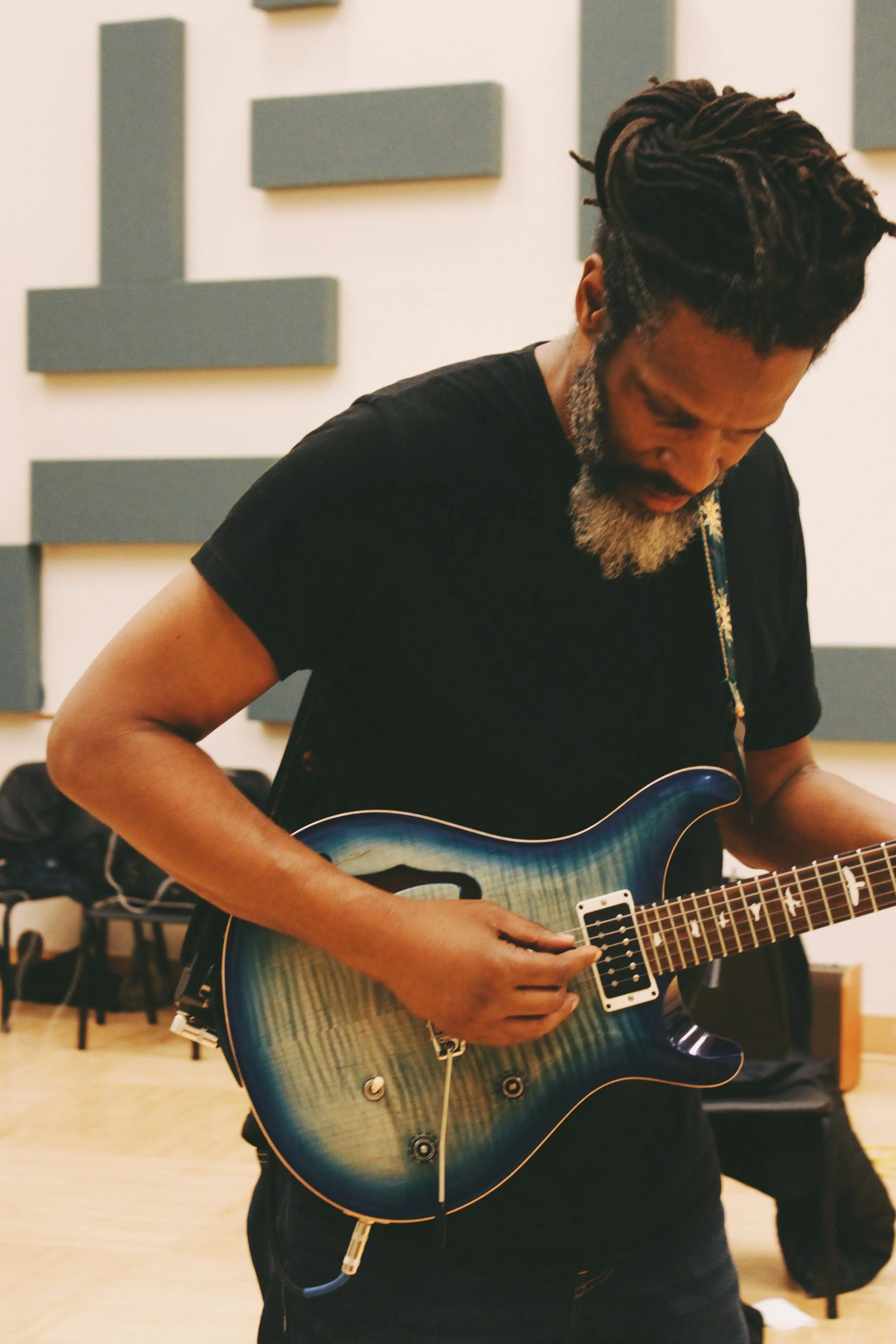 A man with dreadlocks and a beard playing a blue electric guitar in a music rehearsal room.