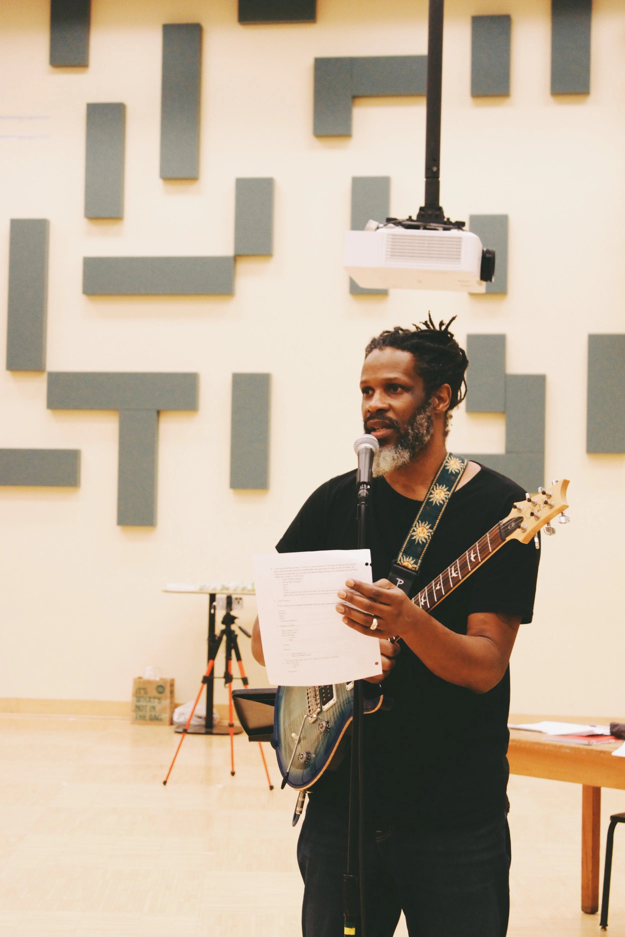 A man with dreadlocks and a beard holds a sheet of paper and a guitar while speaking into a microphone in a music rehearsal space or studio with acoustical panels on the wall.