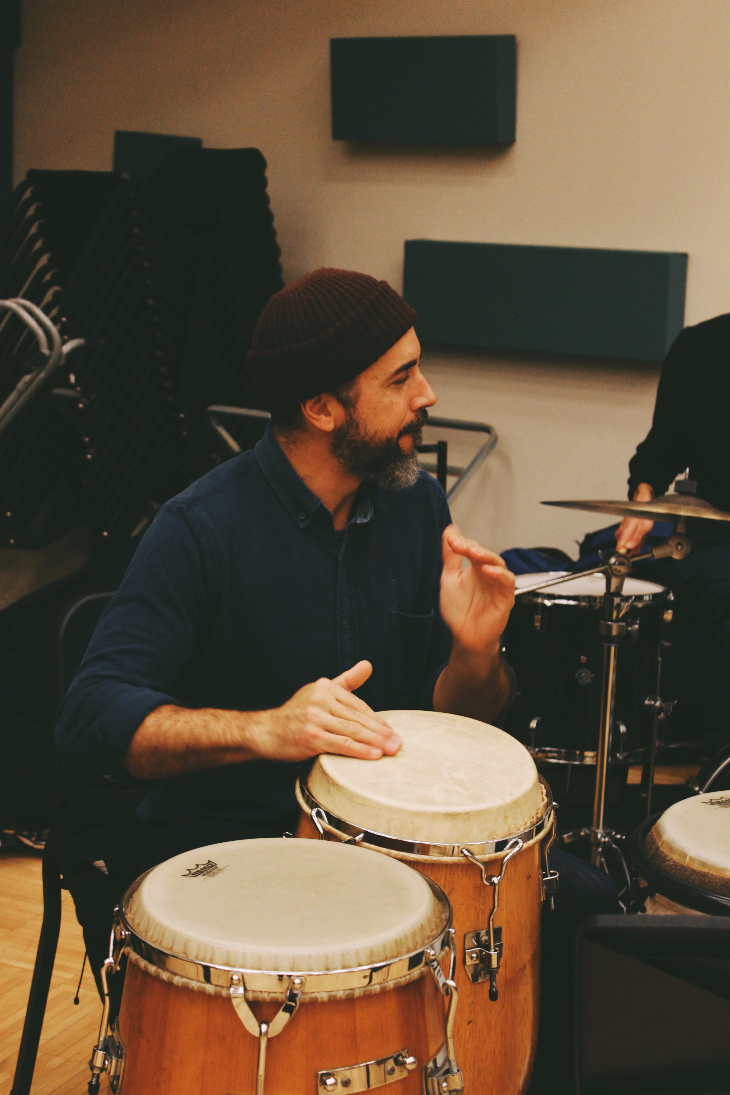 A man with a beard and a maroon beanie plays conga drums indoors.