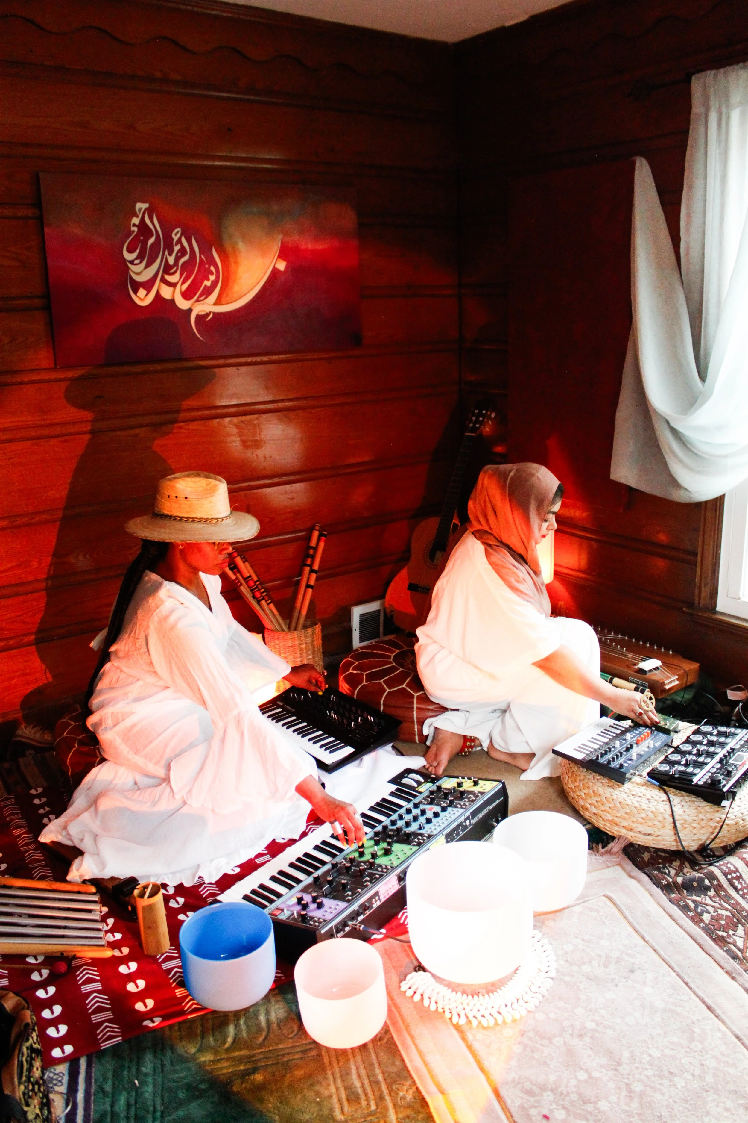 Two women in white dresses and head coverings playing musical instruments, surrounded by colorful bowls and electronic music equipment, in a cozy wooden room with a window and artwork on the wall.