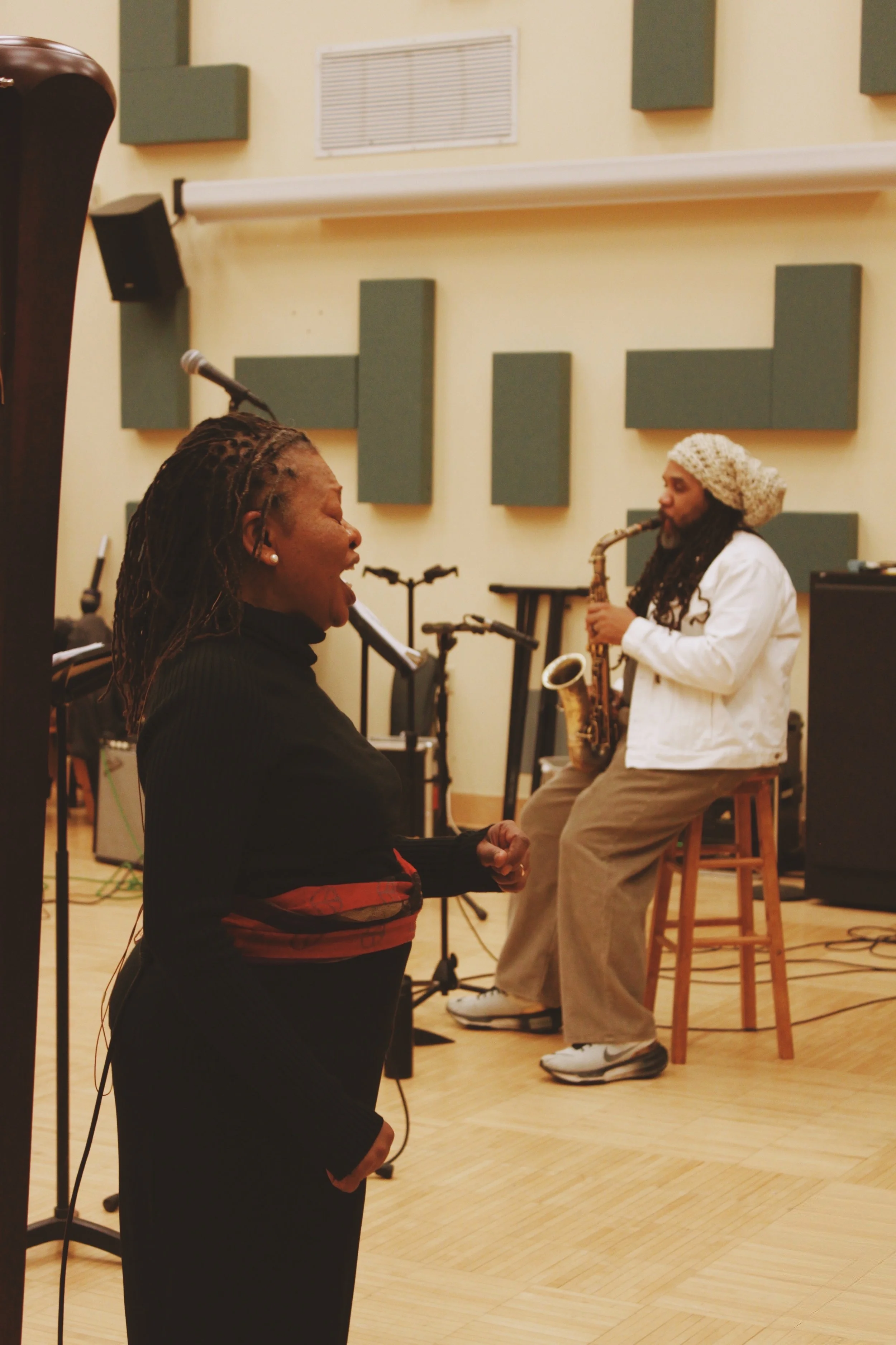 A woman singing passionately in front of a musician playing a saxophone in a recording studio or rehearsal space.