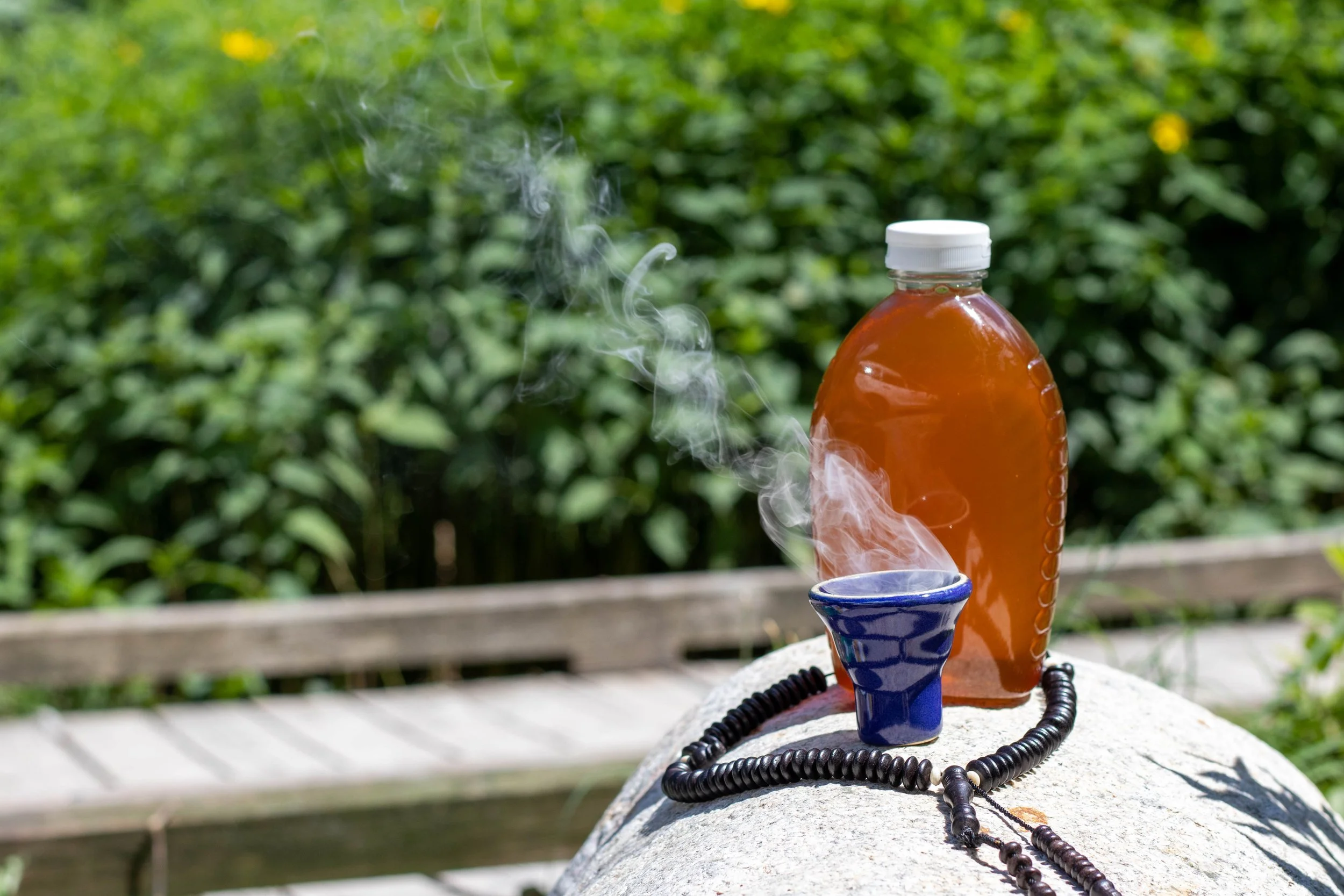 Honey, a blue incense holder with lit incense, and a black prayer beads on a large rock outdoors with green bushes in the background.