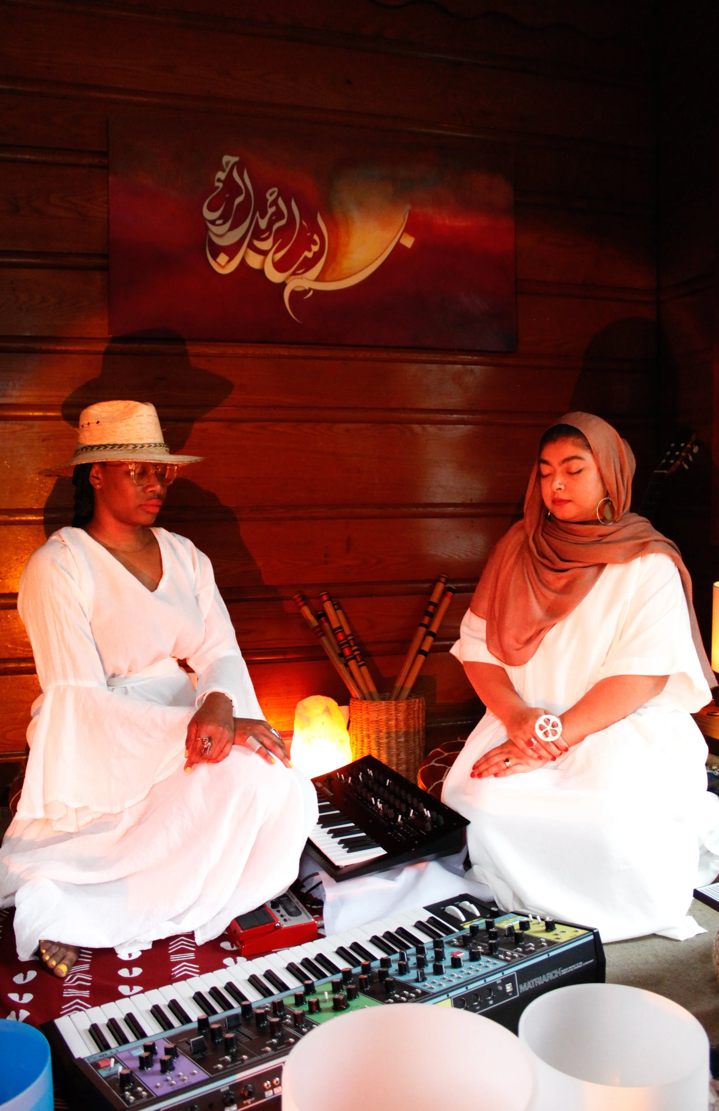 Two women sitting cross-legged with musical instruments on a wooden floor, indoors, in a cozy, warmly lit room.